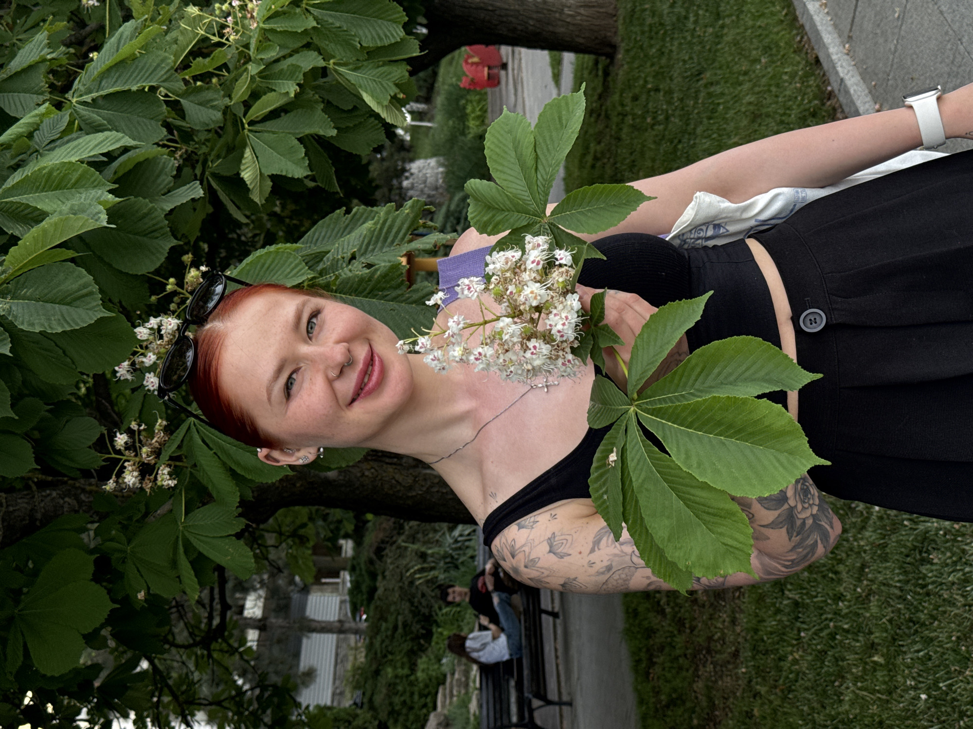 A portrait of a red-haired woman in a park in Rostov-on-Don, Russia. The woman is wearing a black tank top and black pants with a button closure, and has several visible tattoos on her arm and chest. She is holding flowering horse chestnut branches, and sunglasses are perched on her head. In the background, other people are sitting on a bench, and there is a tree and grassy area. The setting appears to be a public park or garden.