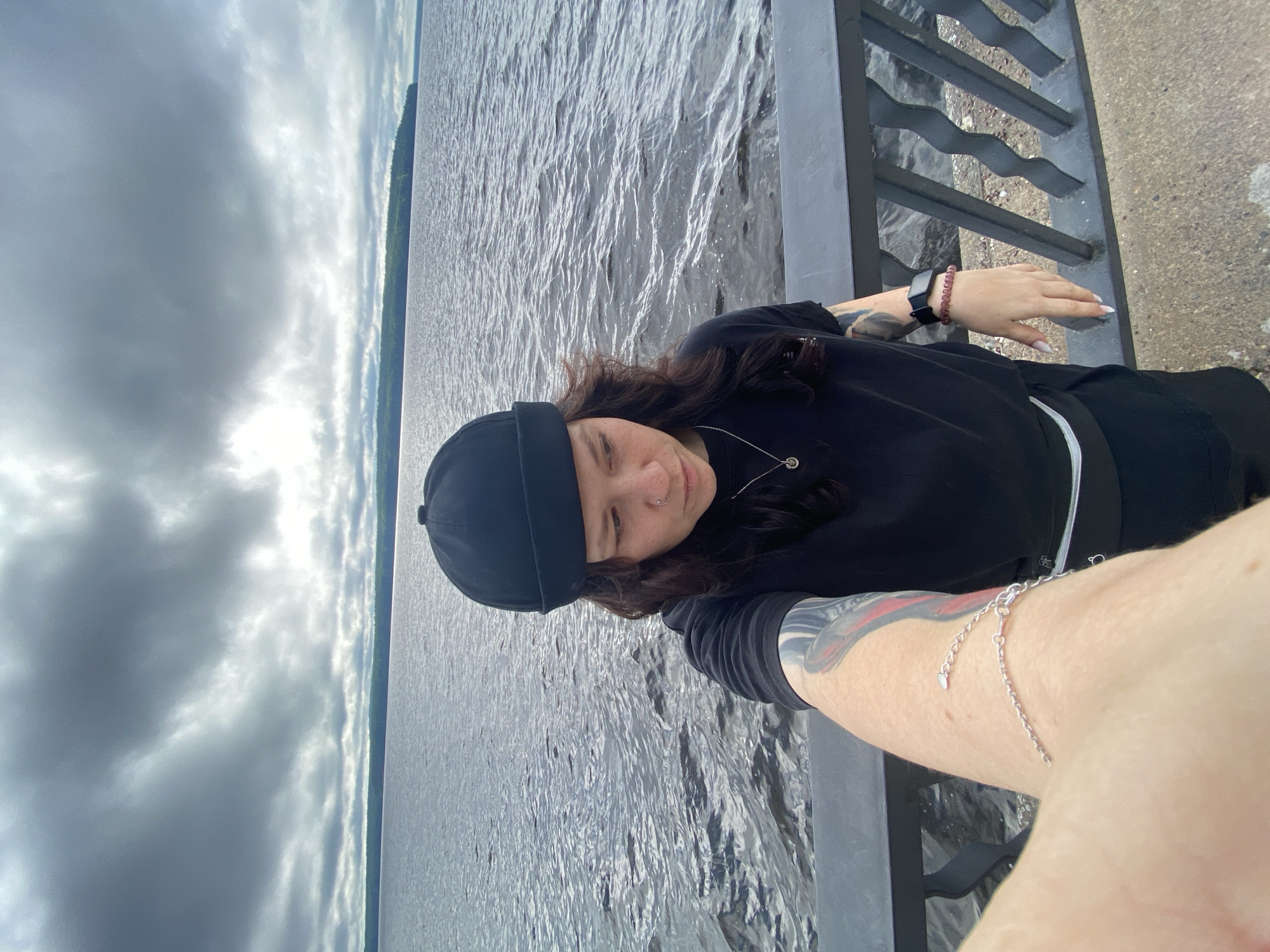 A person stands on a concrete promenade by a large body of water, likely a reservoir. The person is wearing a black beanie, black t-shirt, and black pants. They have tattoos on their left arm and are wearing a silver necklace and a bracelet on their left wrist. A smartwatch is visible on their right wrist. The water is choppy under a cloudy sky. In the distance, a tree-lined shore is visible. The railing of the promenade is in the foreground. The image appears to be a selfie. The location is the embankment of the Kama River in Chaikovsky, Russia.