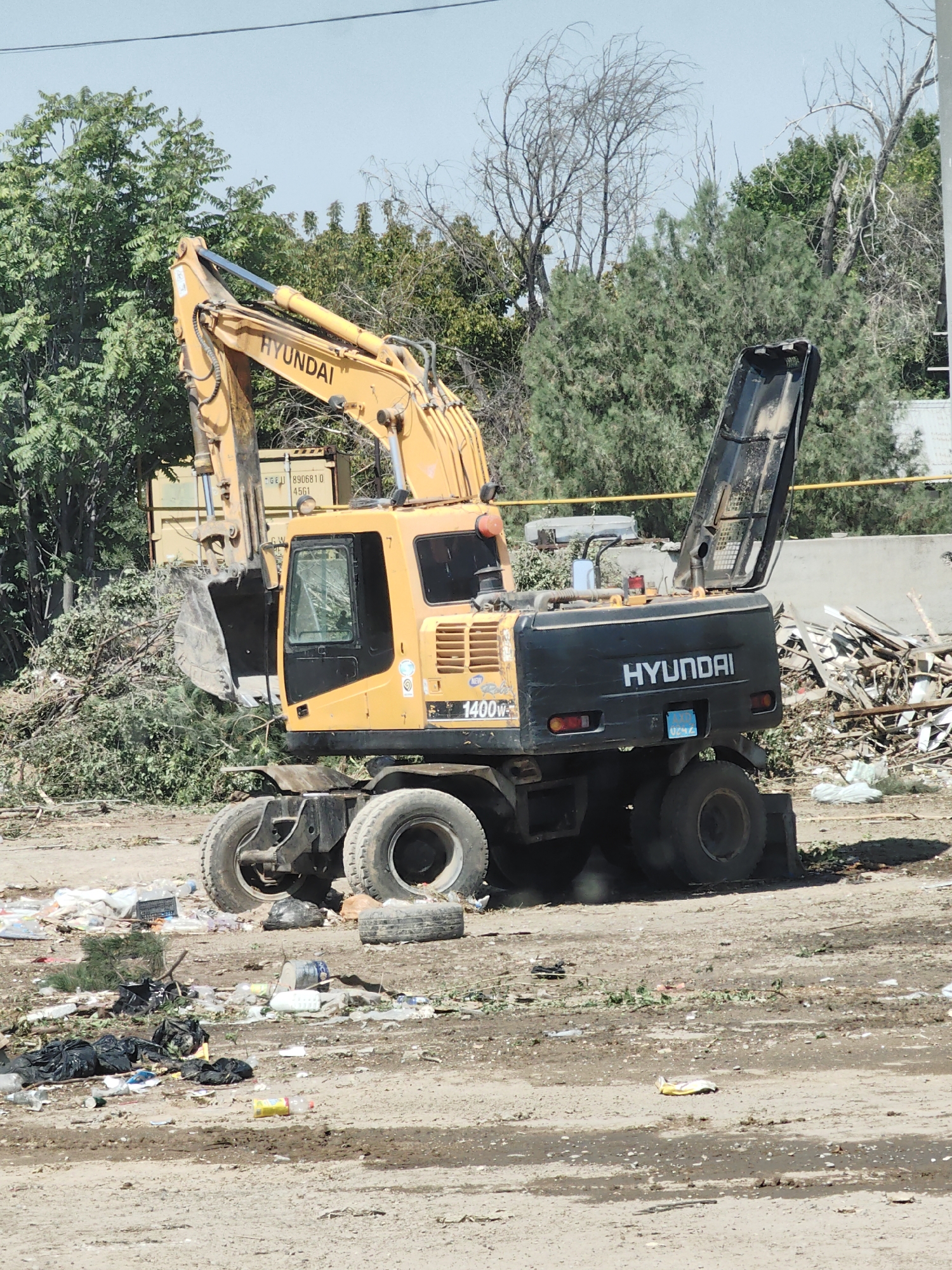 An excavator with "HYUNDAI" branding, identified as a 140W model, is situated in a debris-strewn, outdoor environment. The excavator is yellow and black, with four large tires. It is surrounded by piles of branches, trash, and some discarded building materials. In the background, there are trees and a light blue sky. A shipping container with markings "GE 11 8906810 4561" is visible behind the excavator. The scene suggests a construction or demolition site, or an area where waste is being cleared or dumped. There are no people visible in the image. The location is the city of Shymkent, Kazakhstan.