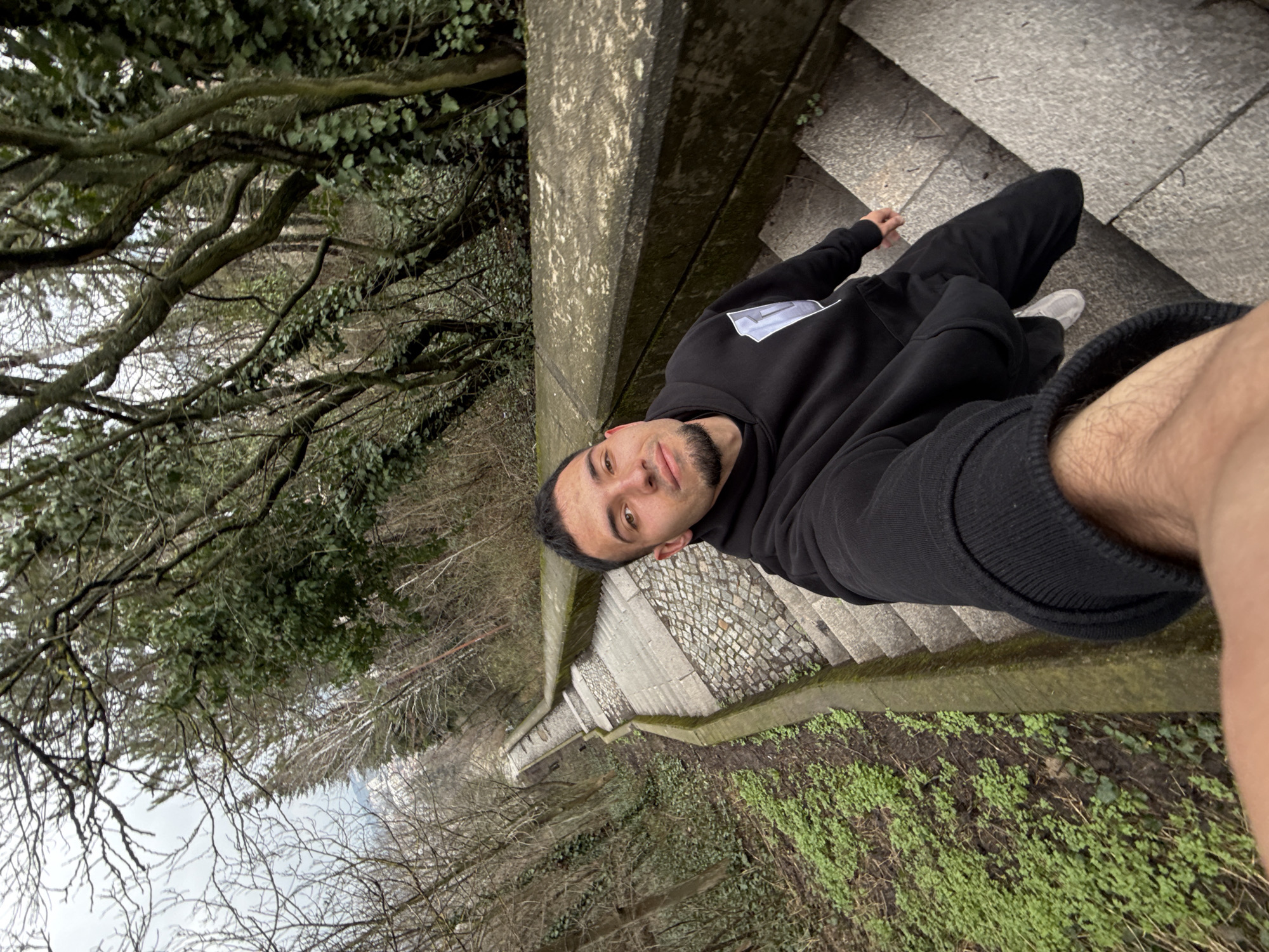A person with brown hair and a trimmed beard is taking a selfie while lying on a set of stone stairs. The stairs are surrounded by trees and foliage. The person is wearing a black hoodie and dark pants. One leg is visible in the foreground. The scene is taken in an outdoor environment.
Based on the unique architecture of the stairs and railings, and the presence of foliage consistent with a park setting, the location is likely the stairs leading down from the Monument to 1300 Years of Bulgaria in Shumen, Bulgaria.