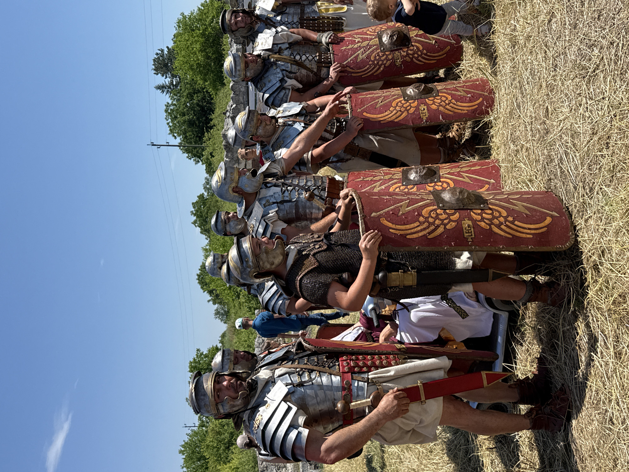 This outdoor image, set in the city of Gigen, Bulgaria, captures a group of approximately ten individuals dressed as Roman legionaries under a clear, bright blue sky on a sunny day. The scene unfolds on a field covered in dry, light-colored straw-like grass.
In the foreground and mid-ground, the majority of the individuals are men attired in authentic-looking Roman military gear. Several wear polished, silver-colored lorica segmentata (segmented plate armor) over light-colored tunics, paired with matching helmets featuring prominent cheek guards. One individual wears lorica hamata (chainmail armor). Each reenactor is equipped with a large, rectangular scutum (shield) predominantly red, adorned with golden-yellow stylized eagle wings and lightning bolts, and featuring a central metallic boss. The letters "LEG" are visible on some shields. Red scabbards for swords are attached to the waists of multiple figures. The reenactors are standing, generally facing forward or slightly to the right, with one individual in the center-right pointing towards a shield.
To the far right, a small child, a toddler, is present, wearing a blue t-shirt and light-colored pants, standing on the grass and looking down. In the mid-ground, behind the reenactors, a white modern golf cart is partially visible, with a person in white attire seated inside.
The background features a line of green deciduous trees. On the left side of the background, a utility pole with overhead power lines is visible. Faint stone structures, consistent with archaeological ruins, can be discerned behind the trees and individuals in the mid-background. The sky above is a uniform clear blue, with one faint, wispy cloud in the upper left quadrant.