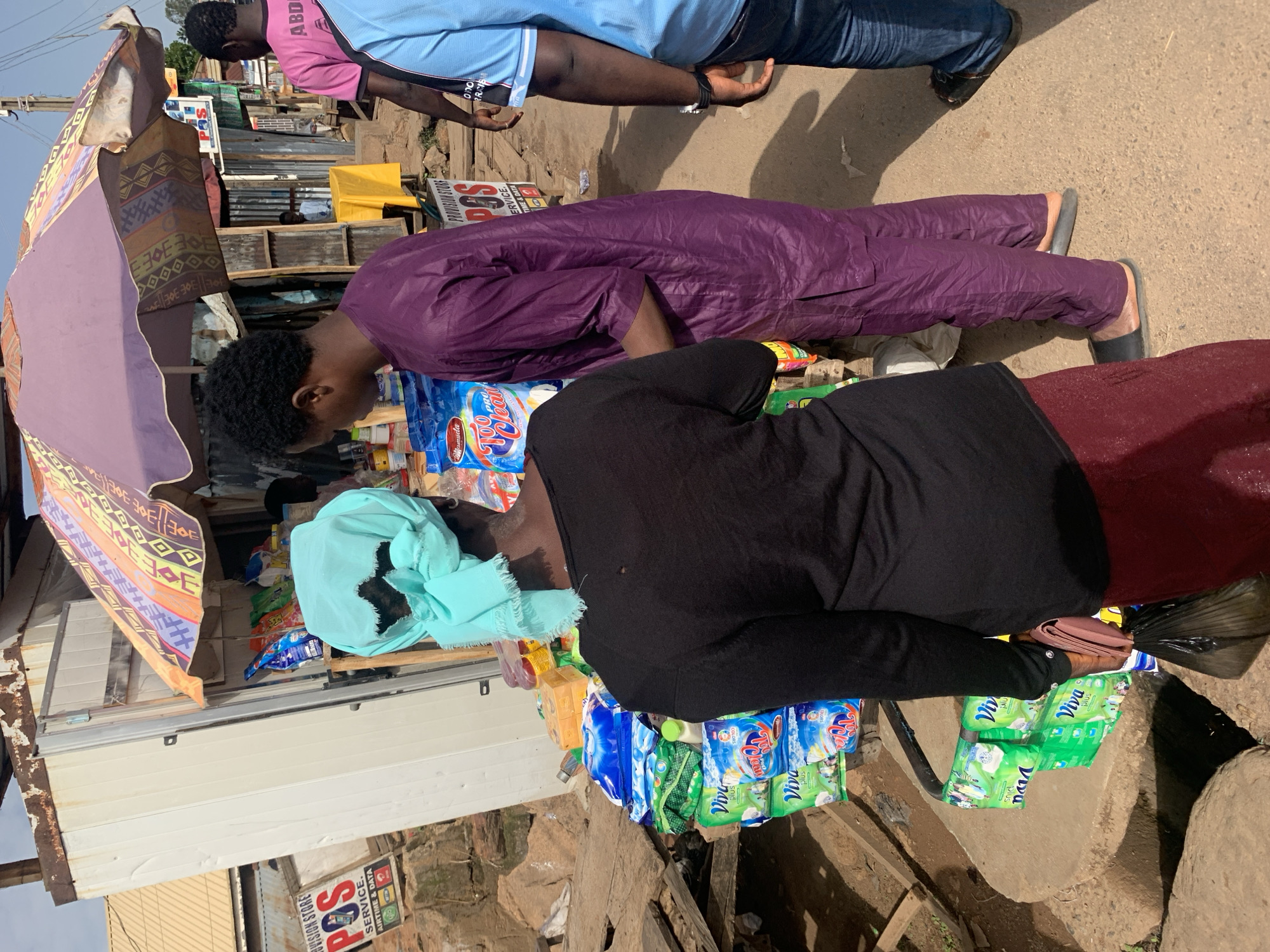 An outdoor scene in Gura Top, Nigeria, features a provision store with goods displayed under a colorful umbrella. A woman wearing a black long-sleeved shirt, a maroon skirt, and a turquoise headscarf stands with her back to the camera, holding a pink purse and a black plastic bag. To her right, a man in a purple tunic and black sandals faces away from the camera. In the background, another man in a pink t-shirt and a person in a blue t-shirt are visible. The ground is dirt, and a white corrugated metal structure, likely part of the store, is present. Several product packages, including "Viva Plus" detergent, are stacked on a table and a concrete structure. A sign for "Provision Store POS Service" is also visible.