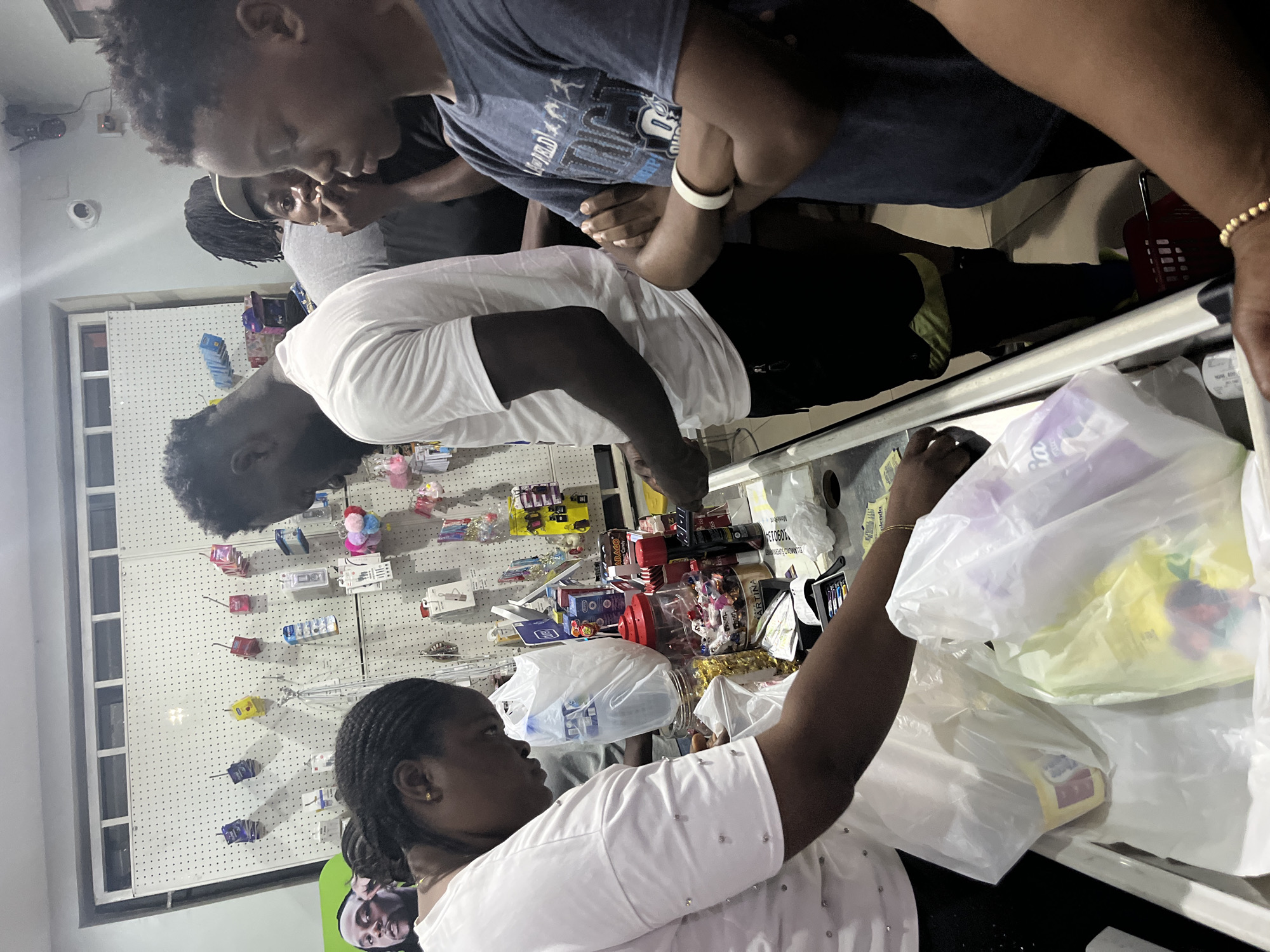 In an indoor setting, several people are gathered around a counter, likely in a store or shop. A woman in a white shirt stands behind the counter, reaching into plastic shopping bags, possibly bagging items. Other people are present, some looking at the items on display, and one wearing a blue shirt. The backdrop includes a wall with various items hanging on it, and there are shelves or displays behind the counter filled with goods. The location is Abuja, Nigeria.