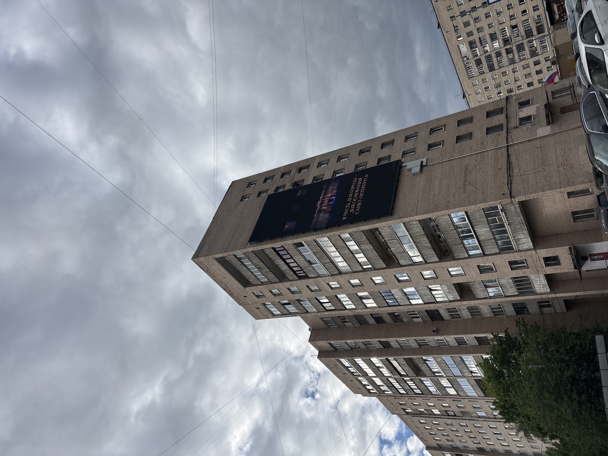 A low-angle, medium shot of two buildings in Saint Petersburg, Russia. The buildings are made of beige brick and are tall. A large, dark sign is attached to one of the buildings. The sign is an advertisement. Power lines are visible against a cloudy sky. Some cars are parked in front of the buildings. A Russian flag is hanging from one of the buildings.