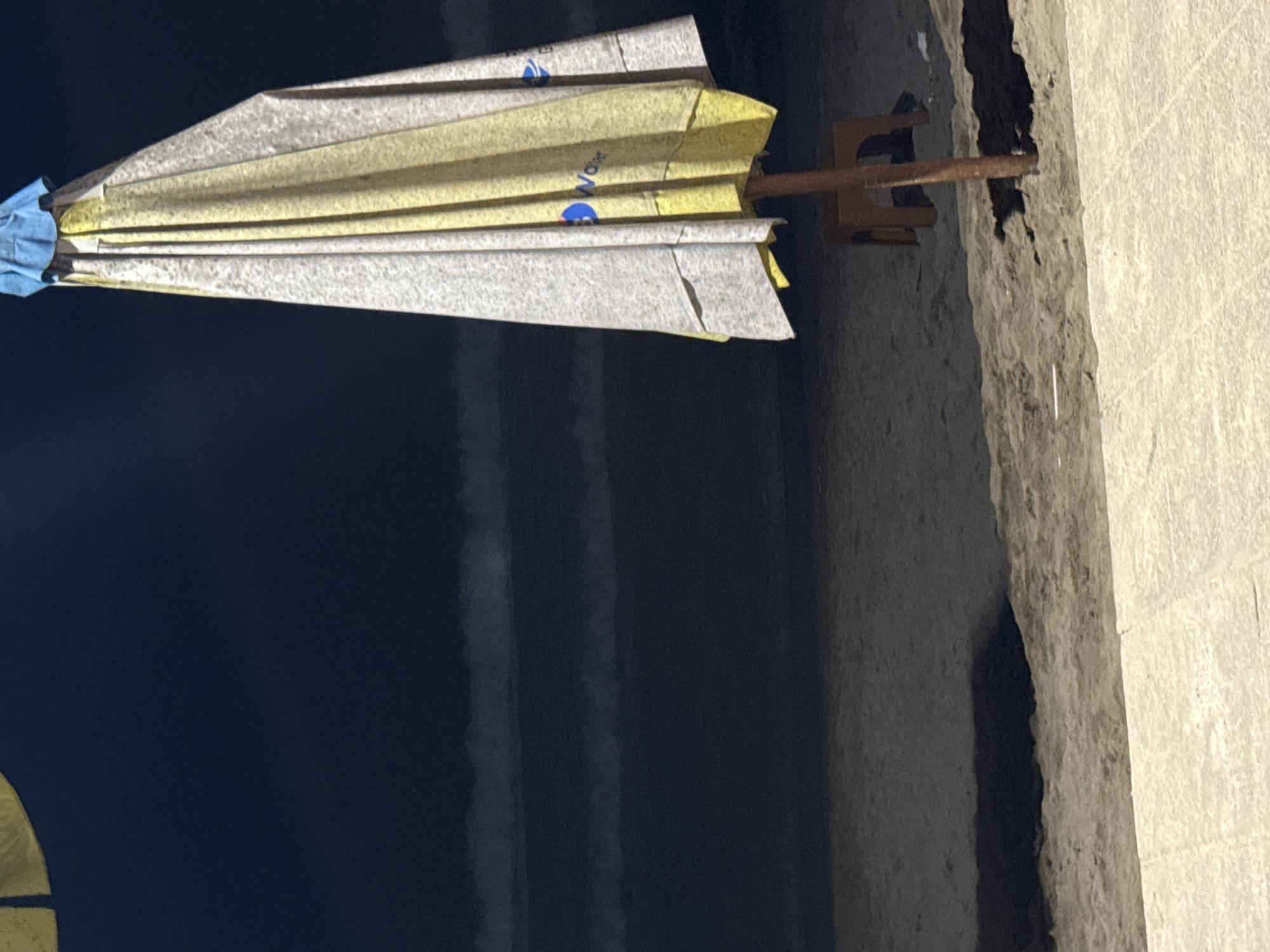 The image, originally rotated, features a collapsed beach or patio umbrella standing upright. The umbrella is predominantly closed, showcasing alternating segments of textured light gray/white and yellow material, possibly paper or thin fabric. Near the top, a section of blue fabric or binding is visible. Several gray segments display the blue text "Water" alongside a blue logo, which resembles a stylized 'W' or a water droplet.
The umbrella is supported by a rusty, dark brown metal stand, characterized by a U-shaped base and a vertical rod. This stand is positioned on a light gray, textured concrete or stone surface, which appears to be a wall or ledge. A dark, irregular crevice or shadow is visible in the concrete behind the stand.
The background to the left of the umbrella is dominated by a vast, dark blue-black expanse, possibly a body of water, a deep shadow, or a dark wall, with faint horizontal lines visible within it. The scene depicts a stationary object in an outdoor setting, likely in Kuta, Indonesia, suggesting the umbrella is unattended. No human activity or specific time of day is discernible beyond general daylight.