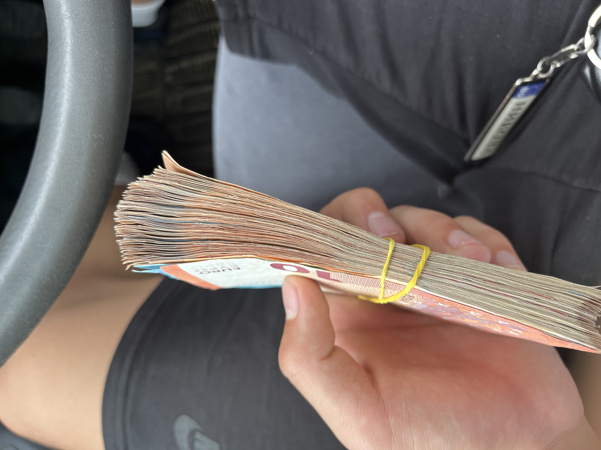 The image shows a close-up view of a person's hand holding a thick stack of Euro banknotes, secured by a bright yellow rubber band. The banknotes are predominantly orange and reddish in color, characteristic of 10 Euro bills, with "EURO", "ΕΥΡΩ", and parts of "10" and "BCE" visible on their faces. The person has light-toned skin and visible fingernails.
The setting appears to be inside a vehicle, with a textured, grey steering wheel or similar interior component partially visible on the left side of the frame. The person is wearing dark grey or black clothing, possibly a t-shirt or jacket, over a lighter grey layer, and black shorts or pants are also visible.
On the upper right, a metallic keyring with a rectangular tag is attached to the person's clothing. The text on this tag is blurry and reversed, making it illegible. The lighting suggests it is daytime. The activity is the simple act of holding the bundle of money. The scene takes place in Ivailo, Bulgaria.