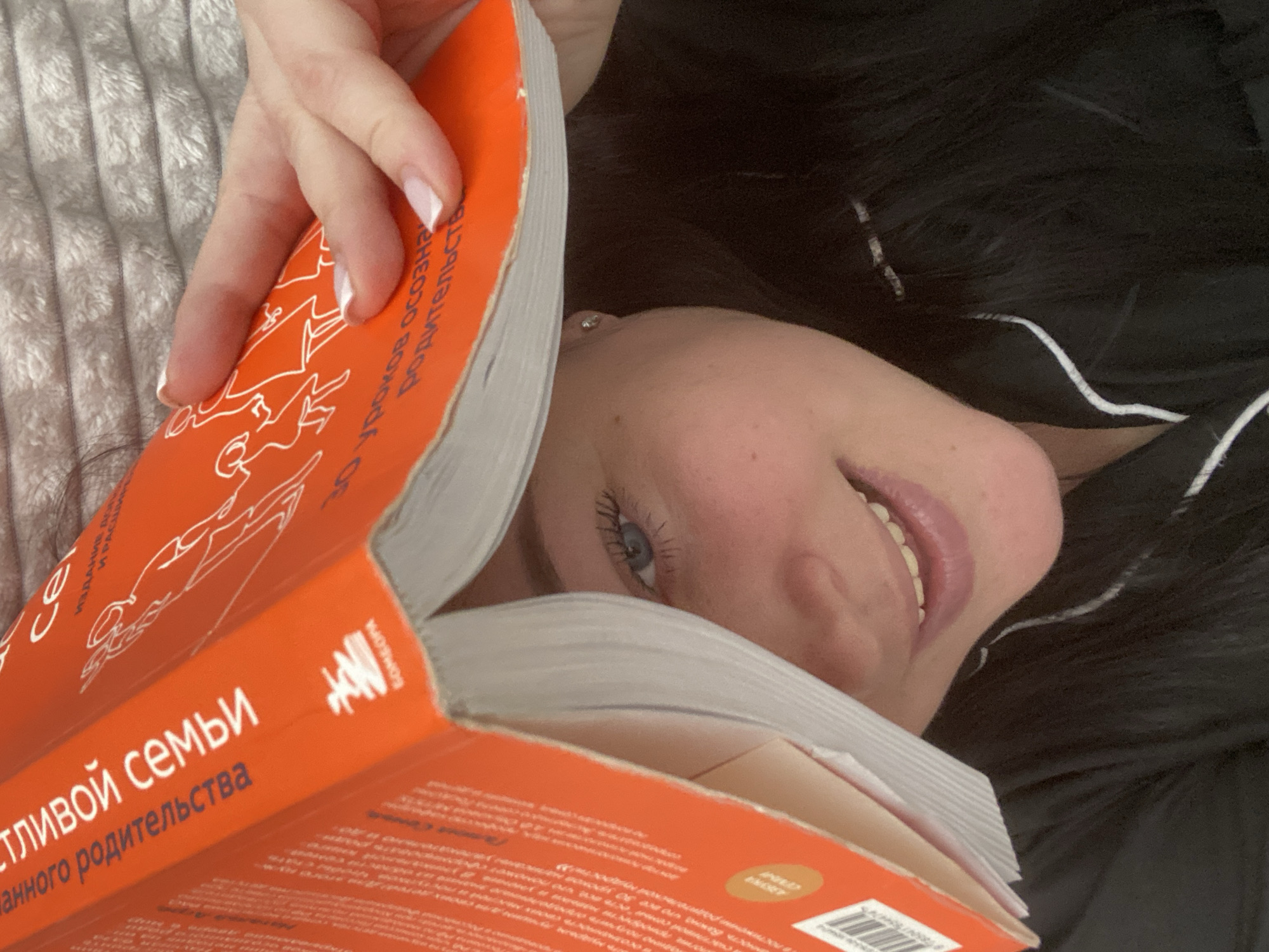 An overhead shot captures a woman lying down indoors, partially obscured by a book she is holding. Her upper body is visible, including her left eye with noticeable blue irises, a slight smile on her lips, and part of her cheek and chin. Her dark hair is spread out on a dark surface, possibly a pillow or blanket, and she appears to be wearing dark clothing with a white stripe, suggesting pajamas. A small, sparkling stud earring is visible in her left ear. Her hand, with nails painted in a French manicure style, holds open a bright orange book.
The book's cover features white line art depicting abstract figures, possibly symbolizing family. Russian text is prominent on the cover, including "СЧАСТЛИВОЙ СЕМЬИ" (Happy Family) and partially visible words like "ДАННОГО РОДИТЕЛЬСТВА" (conscious parenthood), suggesting the book's theme is about family or parenting. The publisher's logo, "МИФ" (MIF), is also visible, along with a small gold circle containing "WORLD BESTSELLER" in English. The woman is resting on what appears to be a soft, textured white or light-colored blanket. Her relaxed posture and gentle smile convey a sense of contentment while reading. Although the image does not provide visual cues for the time of day or the specific location, the context indicates the location is Sochi, Russia.