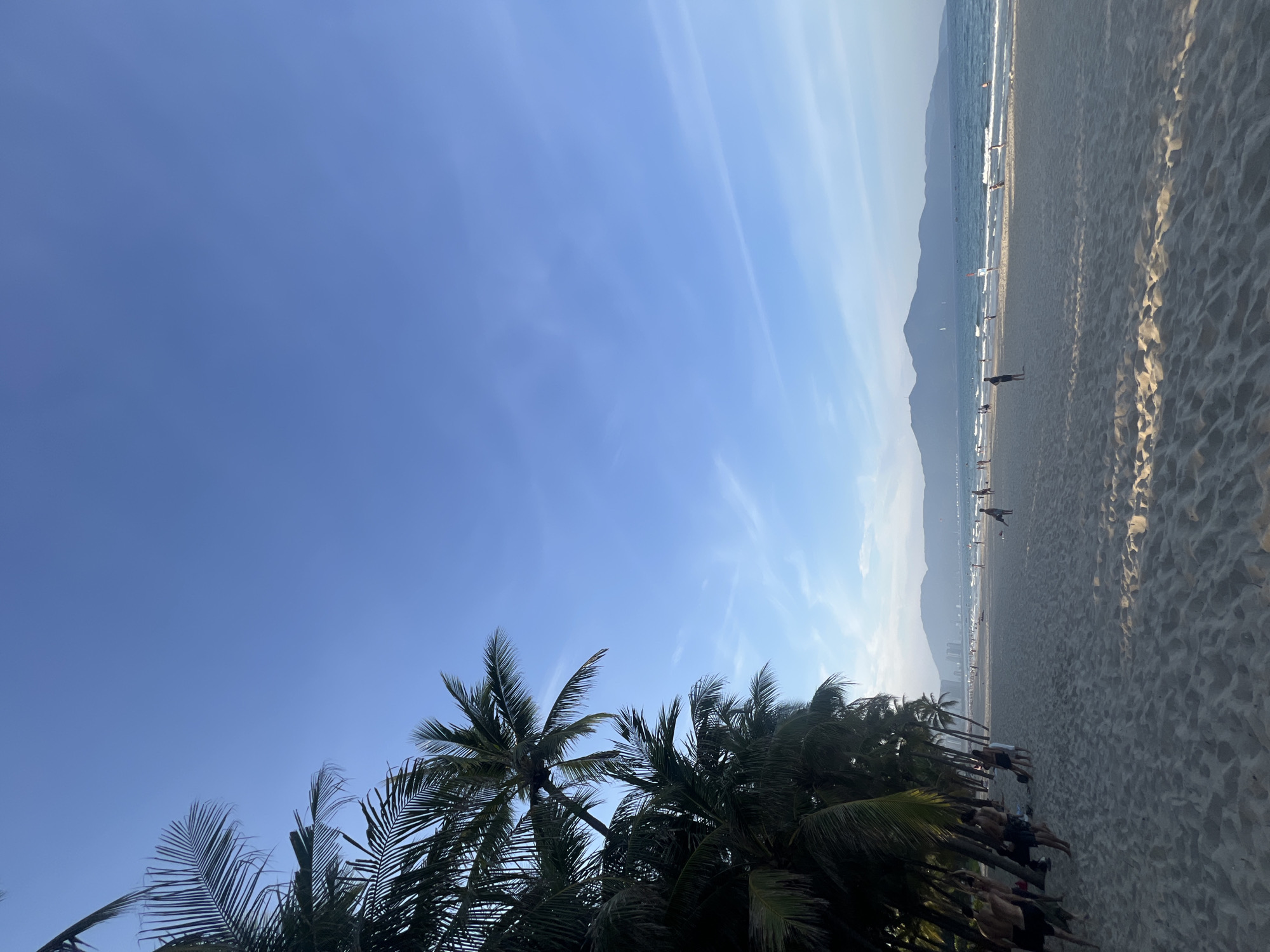 This media file depicts a beach scene in Đà Nẵng, Vietnam. The image is oriented vertically, showcasing a wide expanse of sand stretching towards the ocean. The sky is a clear blue with faint, wispy clouds, suggesting a pleasant day. In the foreground, the fronds of several palm trees are silhouetted against the bright sky, adding a tropical element to the scene.
Further down the beach, a scattering of people are visible. Some are walking along the shoreline, while others appear to be engaged in activities near the water's edge. A few individuals are also seen in the distance, their forms small against the vastness of the beach and the sea. The ocean itself is calm, with gentle waves lapping at the shore.
In the background, a range of mountains forms a hazy silhouette against the horizon, characteristic of the coastal landscape of Đà Nẵng. The lighting suggests it is likely late afternoon or early morning, with the sun casting long shadows across the sand. The overall mood is serene and relaxed, capturing the essence of a day spent at the beach. There is no visible text in the image.