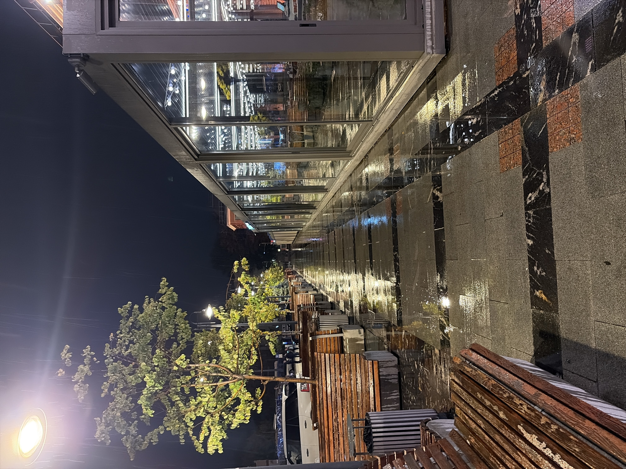 The image depicts a wet pedestrian walkway at night in Moscow, Russia, illuminated by streetlights and interior lights from a glass-enclosed building. Rain is visible falling. The walkway is paved with dark gray and black granite tiles, some of which have reddish-brown square inlays. Wooden benches with metal frames line the left side of the walkway, some with trash receptacles nearby. A white SUV is parked on the other side of the benches. The glass-enclosed building on the right side of the walkway reflects the lights and trees. The scene conveys a quiet, possibly deserted, urban atmosphere during a rainy night.