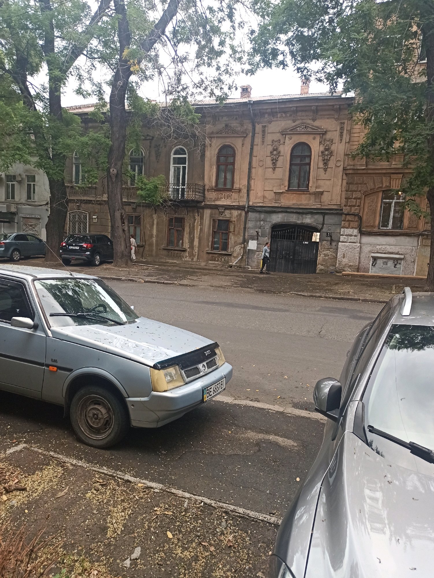 In the city of Odesa, Ukraine, an overcast day captures a street scene with parked cars and an older building. In the foreground, the front section of a light blue car is visible, parked on a paved surface with fallen leaves. To its right, the hood and side mirror of a silver car are also in view. Beyond the cars, a two-story building with ornate architectural details and arched windows stands. A dark metal gate is visible in the center of the building's facade. A person in a light-colored jacket and dark pants is walking away from the gate towards the left side of the frame. A dark SUV is parked further down the street, to the left of the building, and another car is partially visible to its left. Mature trees with green foliage line the street, casting shadows on the pavement and the building.