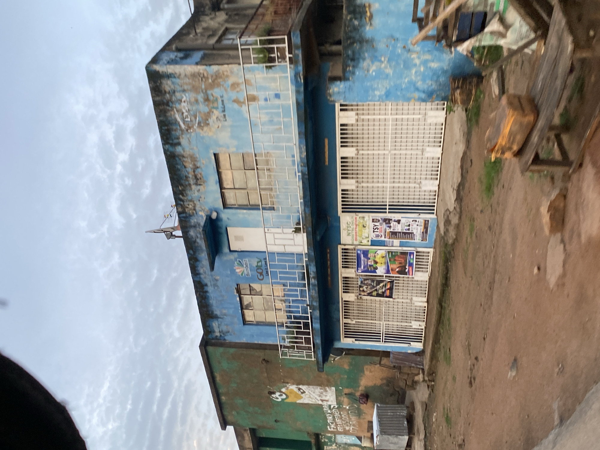 The image depicts a two-story building with peeling blue paint on the upper floor and green paint on the lower left portion. The building is located on a dirt road in what appears to be a commercial area in Gwong, Nigeria. Several businesses are visible on the ground floor, indicated by signs and posters on the metal grates covering the storefronts. These include signs for "MultiChoice" and "GOtv," suggesting a telecommunications or media-related service. There are also posters for "NYSC" and "TSY," which are likely related to national youth service and another service or organization.
On the left side of the building, there's a mural of a person in athletic wear and some text that reads "FACTORY UP... ERT NEEDED... 55,000... 070311100." A corrugated metal bin is situated to the left of the entrance. The upper floor features a balcony with white railings and windows. The sky above is overcast with scattered clouds. In the foreground, the ground is uneven and dusty, with scattered debris, including a wooden bench with a piece of wood on it, and some stacked lumber. The overall scene suggests a somewhat dilapidated but active commercial street.