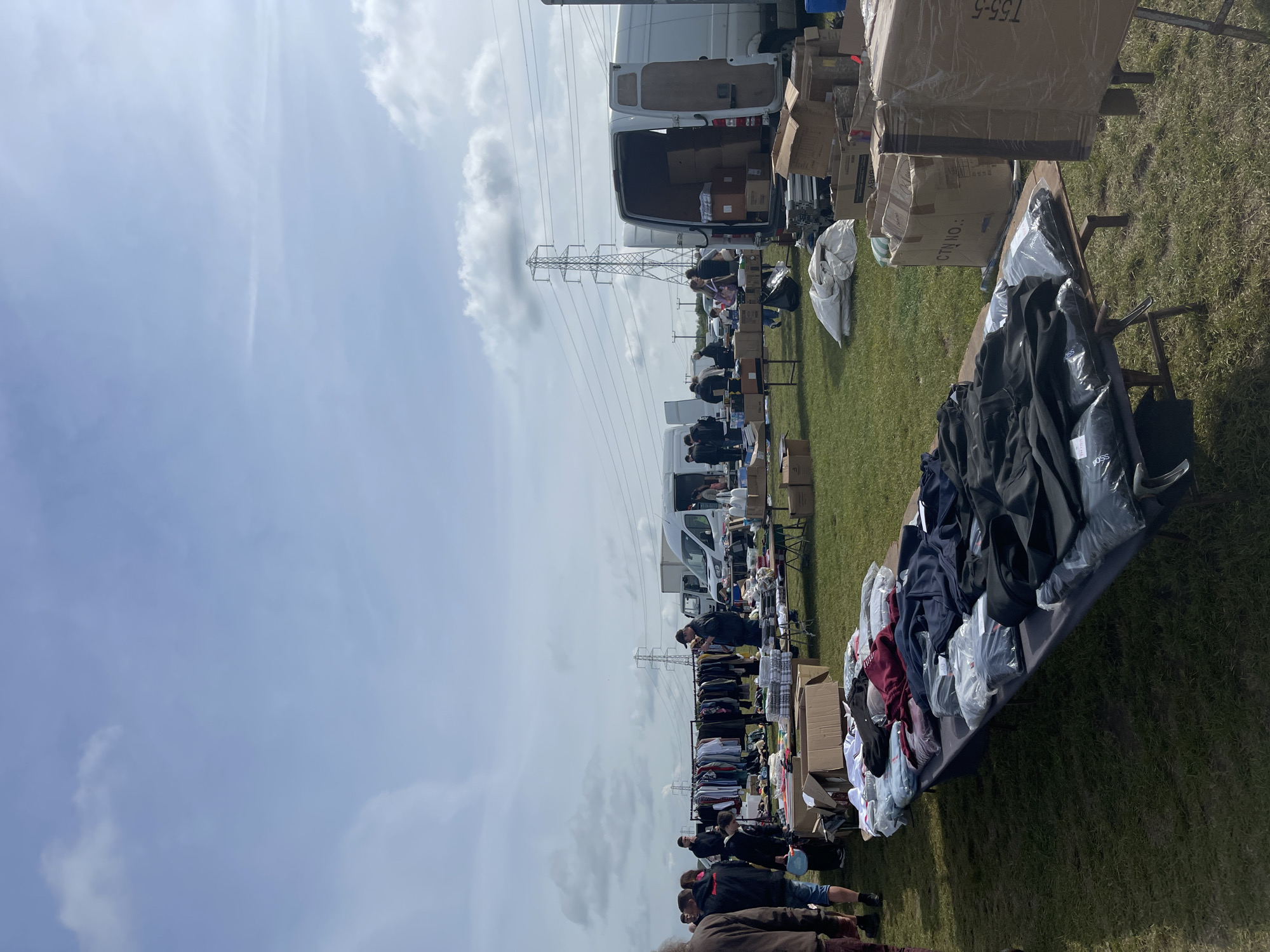 The image shows an outdoor market in Sevenoaks, United Kingdom. Several white vans are parked alongside tables displaying merchandise, primarily clothing. People are browsing the items on the tables. Cardboard boxes and bags are also visible. In the background, there is an electrical transmission tower. The sky is mostly cloudy. The setting appears to be a grassy field.