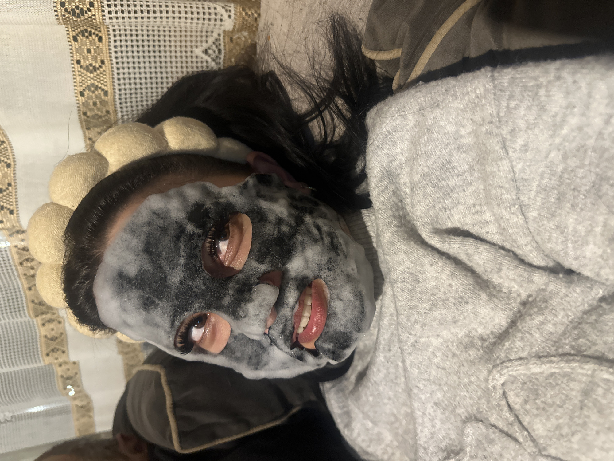 A person is reclining, facing the camera. They have black hair and are wearing a white headband. Their face is covered in what appears to be a bubble face mask. Their eyes are open and they are partially smiling. A gray garment is visible next to them. There is a patterned cloth in the background. The location and city of Chubra, Bulgaria are not identifiable from the image.
