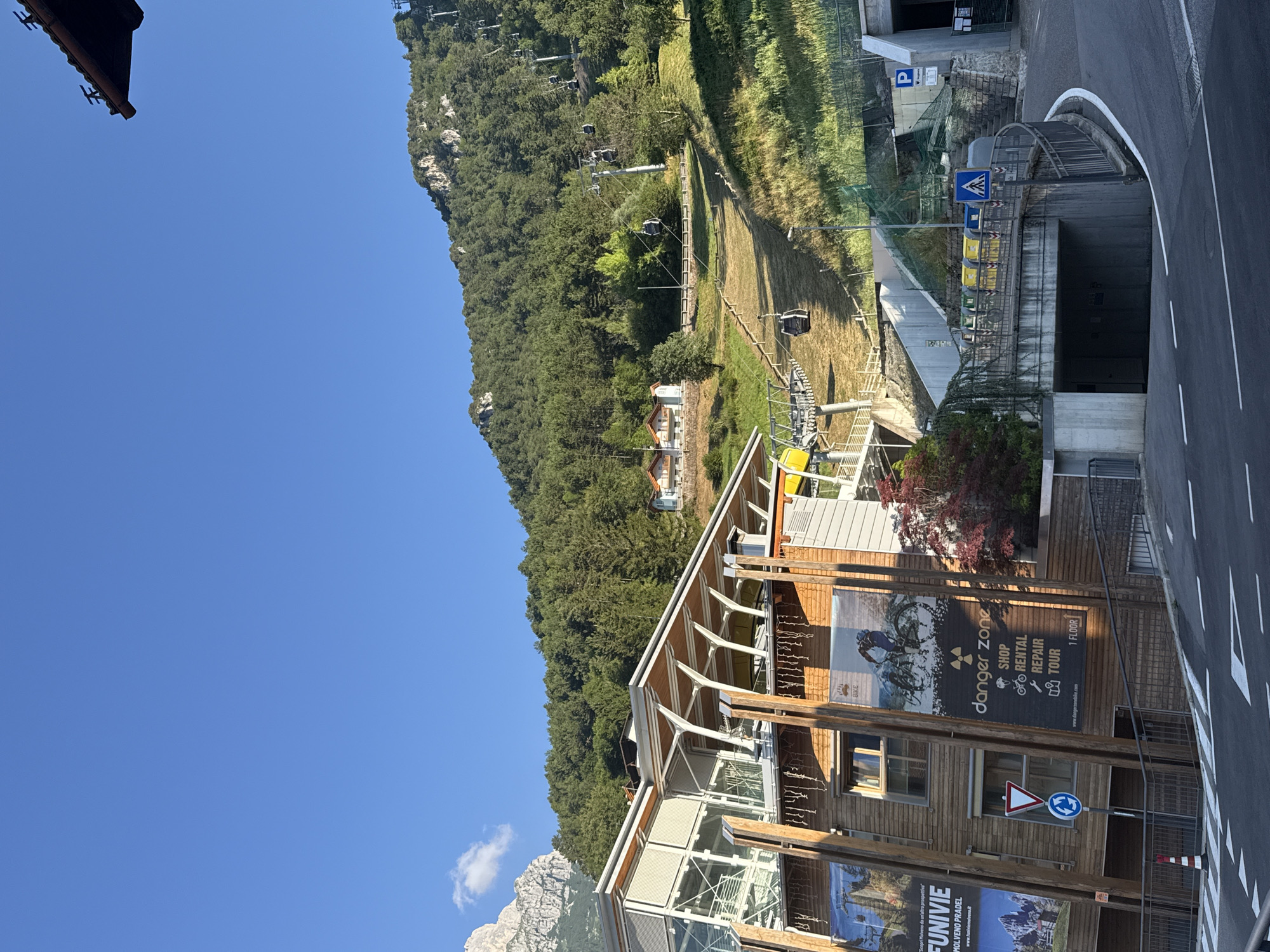 The image depicts a sunny day in Molveno, Italy, with a clear blue sky dominating the upper portion. In the foreground, a modern wooden building with large glass windows and a wooden-beam roof is visible. Large advertisements are attached to the building's exterior, one featuring a mountain landscape and the words "FUNIVIE MOLVENO PRADEL," suggesting it is a cable car station. Another advertisement showcases a mountain biker and the text "danger zone SHOP RENTAL REPAIR TOUR." A traffic sign indicating a roundabout and a warning sign are also present.
Behind the building, a steep, grassy slope rises towards a densely forested mountainside. A cable car system with multiple cabins isAscending the slope, with some cabins visible against the green backdrop. In the distance, two small houses with red roofs are nestled among the trees on the mountainside.
To the right of the image, a road curves towards a concrete tunnel entrance. A parking sign and a pedestrian crossing sign are mounted on a concrete wall. Several colorful recycling bins are lined up along a fenced area. The overall scene suggests a scenic mountain resort area.