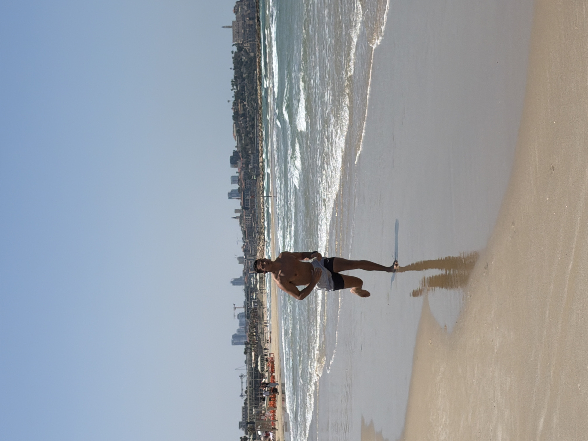 A man is walking barefoot on a beach in Tel Aviv, Israel. The ocean is to his right and a row of buildings and trees is visible in the background. The sky is clear and blue. The man is wearing shorts and is shirtless. His shadow is visible on the sand.