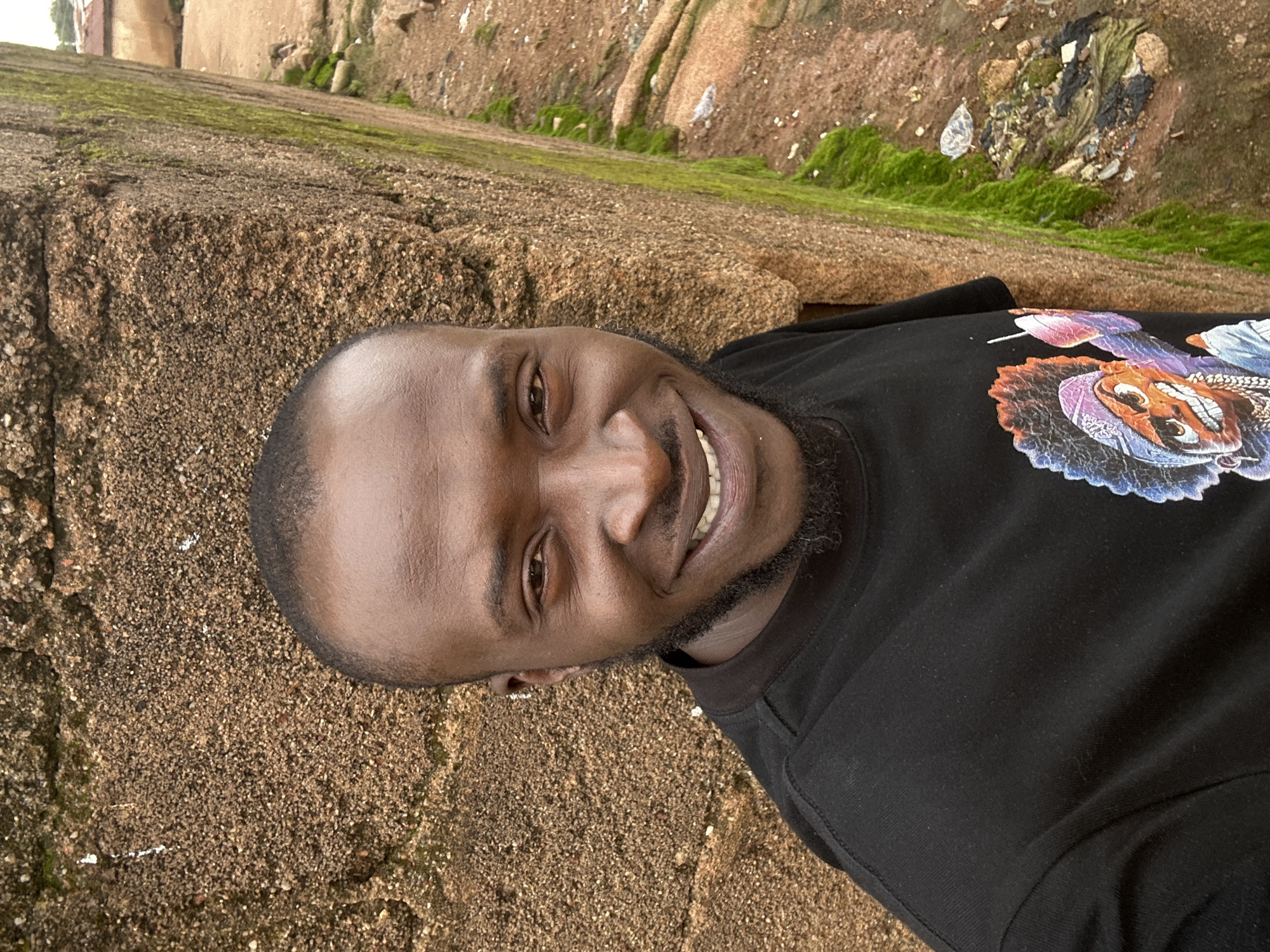 A man with dark skin and short hair, wearing a black t-shirt with a colorful cartoon graphic, stands in front of a textured, sandy-colored wall. The wall appears to be part of an outdoor structure, possibly a building or a retaining wall. To the right of the wall, a patch of vibrant green moss grows along its edge, and further to the right, the ground is visible, covered with dirt, small rocks, and some scattered debris. The background is slightly blurred, showing more of the wall and what appears to be a residential area with some buildings. The scene suggests an outdoor setting in a possibly rustic or less developed part of a city. The man is looking directly at the camera with a slight smile, and his face is well-lit. There are no other people visible in the image. The overall impression is of a casual outdoor portrait taken in Jos, Nigeria.