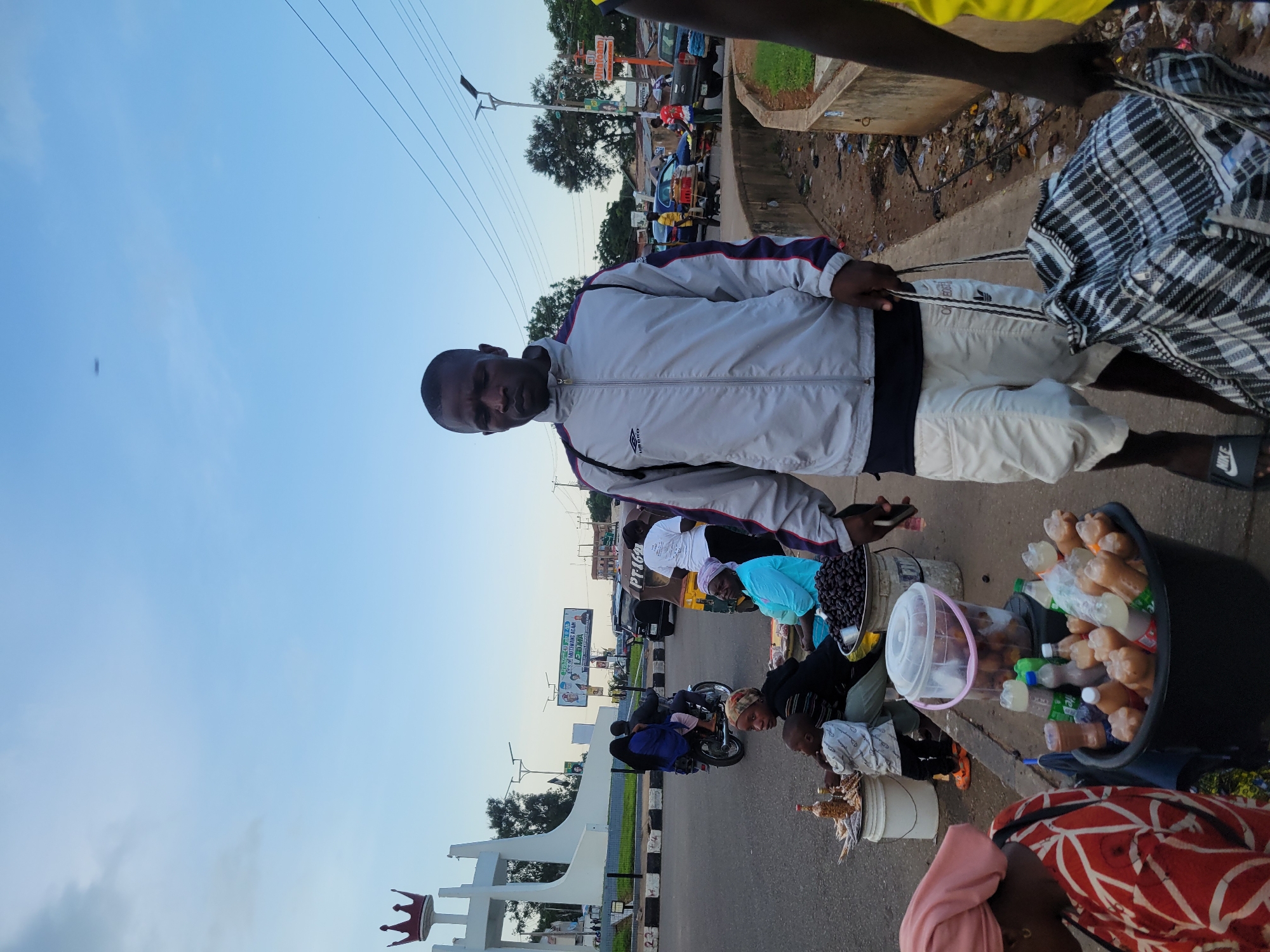 The image depicts a street scene in Barakin Lerawi, Nigeria. In the foreground, a man walks towards the camera, carrying a large checkered bag. He is wearing a light-colored jacket with dark stripes on the sleeves and white shorts. Behind him, to the left, a woman in a red and white patterned top and a headscarf is visible. A large black bucket filled with bottles of beverage is in front of her. Further to the left, a young child in a white shirt stands near a bucket of snacks. Several other individuals are seated and standing near makeshift stalls selling goods. In the mid-ground, traffic moves along a road, including a motorcycle carrying two passengers and an auto-rickshaw. A large white monument with a red crown on top is visible in the background, along with trees and buildings. The sky is clear and blue, suggesting it is daytime. The overall scene suggests a bustling marketplace environment.
