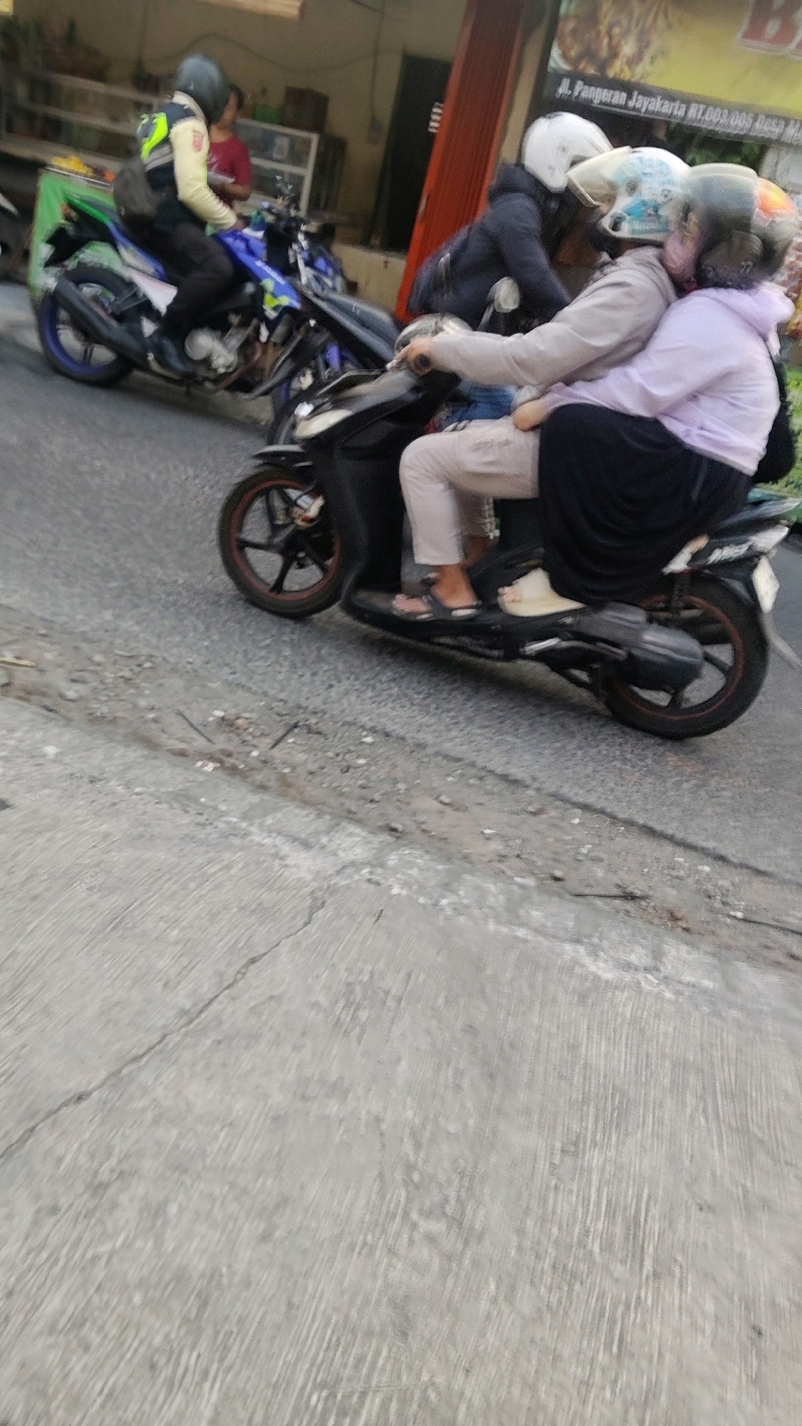 In Mekarmukti, Indonesia, three individuals are riding on a black motorcycle. The rider in the front, wearing a dark jacket and a white helmet, is partially obscured. The middle passenger, wearing a light-colored jacket and a helmet with a blue and white pattern, is positioned behind the rider and appears to be embracing the rear passenger. The rear passenger, wearing a light purple hoodie and a dark hijab, is seated behind the middle passenger, also embracing them. The motorcycle has a blue and yellow design on its side. The scene takes place on a street with a rough asphalt surface in the foreground and a sidewalk with some debris. Buildings with shop fronts and red shutters are visible in the background.