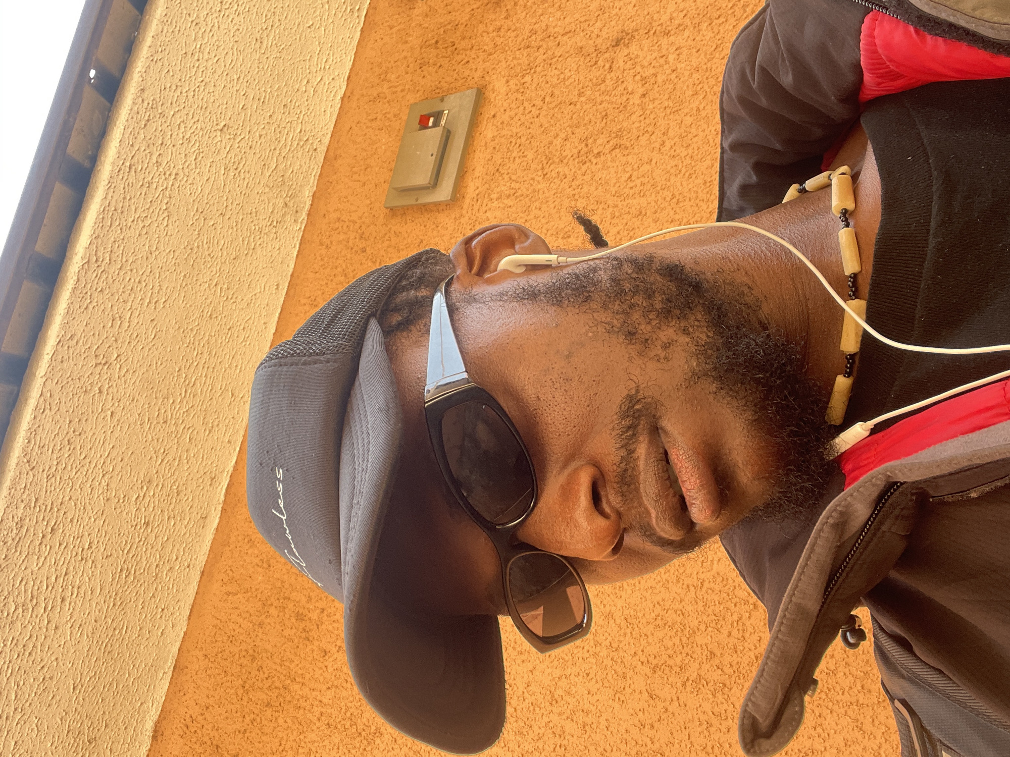 The image is a close-up, low-angle shot of a Black man wearing a grey baseball cap, sunglasses, and a beaded necklace. He also has earbuds in. The man's face is partially visible, with a beard and mustache. He is looking down and to the left.
The man is wearing a dark jacket with red and grey accents. The background is a textured orange wall, possibly stucco. There is a small electrical box mounted on the wall behind him. The lighting suggests it is daytime, and the bright lighting and shadows indicate it might be sunny.
The baseball cap appears to have some white text on the side, though it is partially obscured and difficult to read clearly. The beads of the necklace are light brown and black. The scene appears to be outdoors, possibly in Ban, Nigeria, given the location context. The man's expression is neutral.