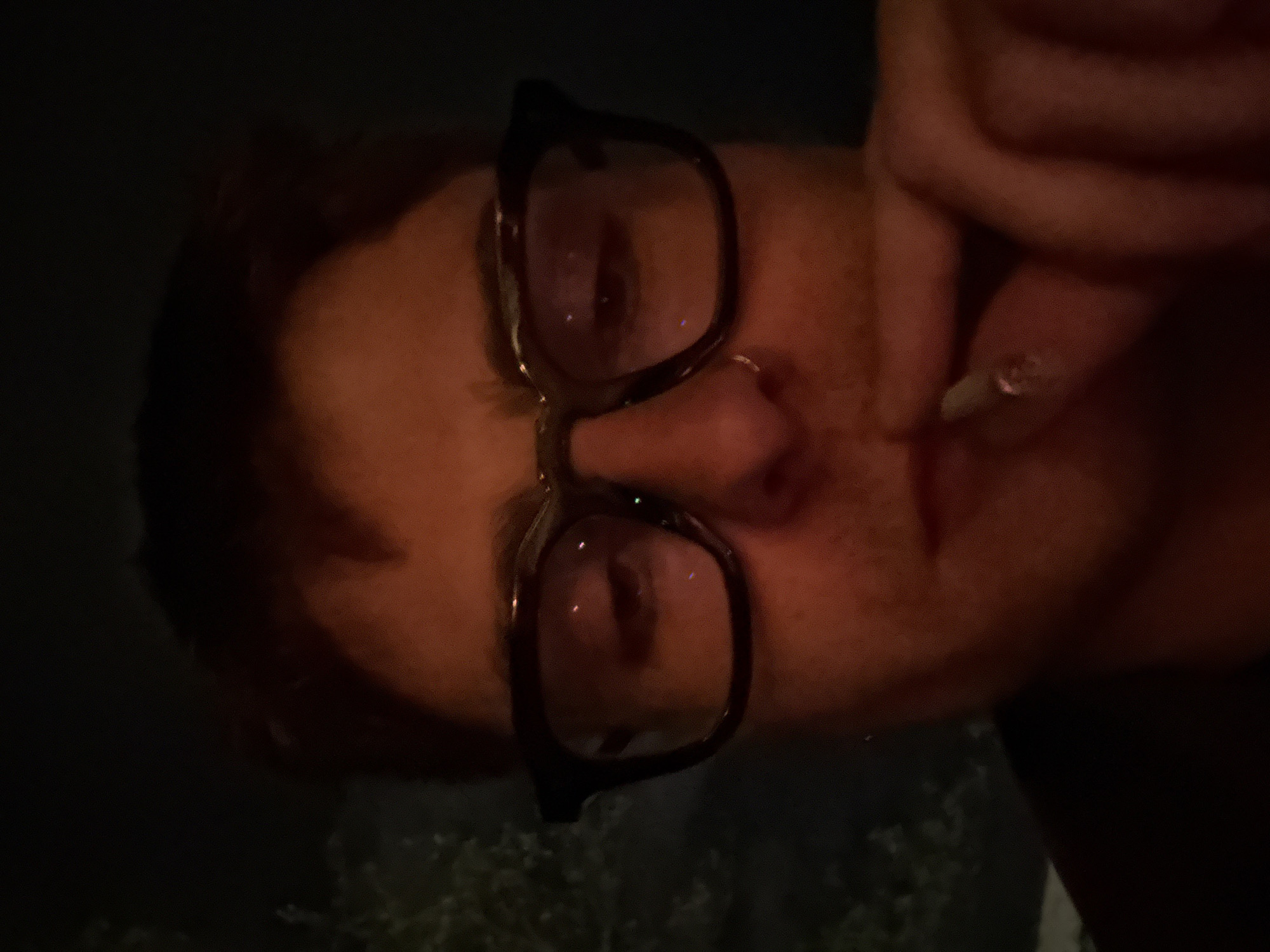 A close-up, low-angle shot shows a person wearing dark-framed glasses and a nose ring. The person is holding a lit cigarette to their lips. The background is dark, possibly night, with faint lights suggesting trees or foliage. It's impossible to determine the exact location of London from this image alone.