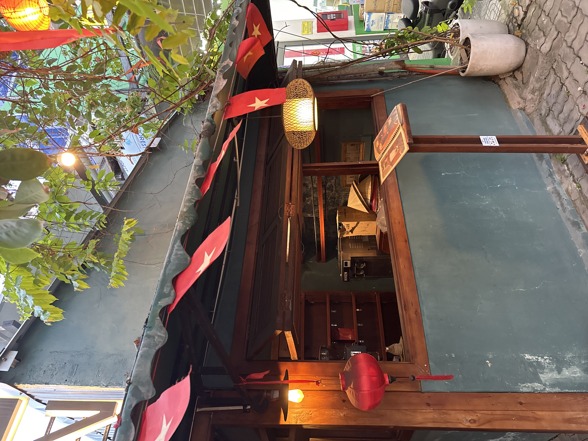 The image depicts the exterior of a small establishment in Da Nang, Vietnam, likely a food vendor or cafe. Red Vietnamese flags are strung along the awning of the building, which is painted a muted teal color. A red lantern hangs from a wooden beam on the left side of the structure. Inside the open-air counter area, a coffee machine and shelving are visible. A wooden menu stand is positioned on the sidewalk in front of the counter. Two large, light-colored pots containing plants are placed to the right of the establishment. The ground is paved with interlocking concrete blocks. The lighting suggests it is late afternoon or early evening. There are no people visible in the frame.