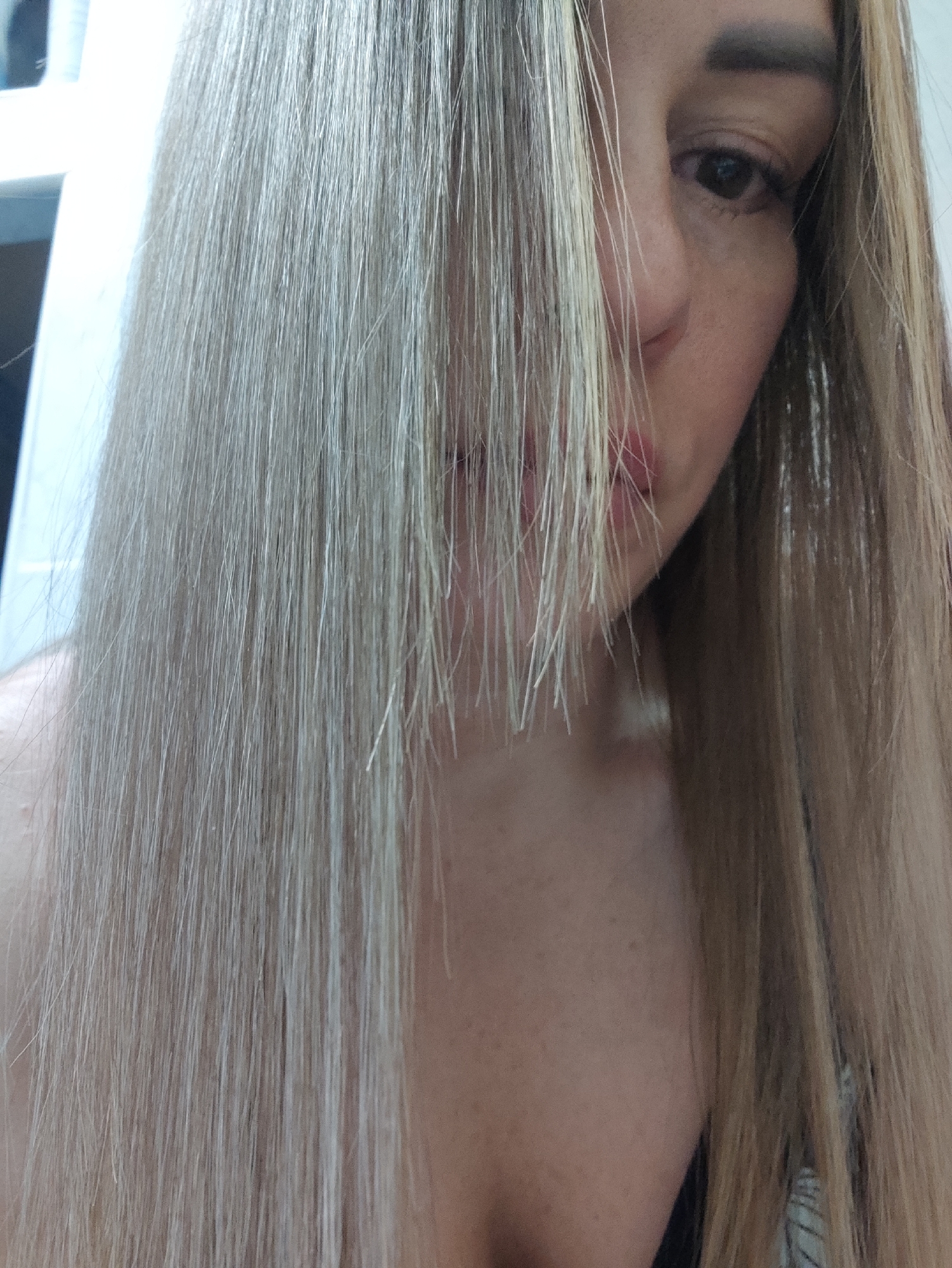 The image is a close-up, portrait-style photograph of a person's face and hair, with their blonde hair obscuring part of their face. The person's left eye, eyebrow, and a portion of their nose and lips are visible. The background is out of focus and appears to be an indoor setting, possibly a room with a white wall and some shelves or furniture. There are no clear indicators of the specific location within Almaty, Kazakhstan, or any specific objects or actions that would place it there. The lighting suggests it is daytime.