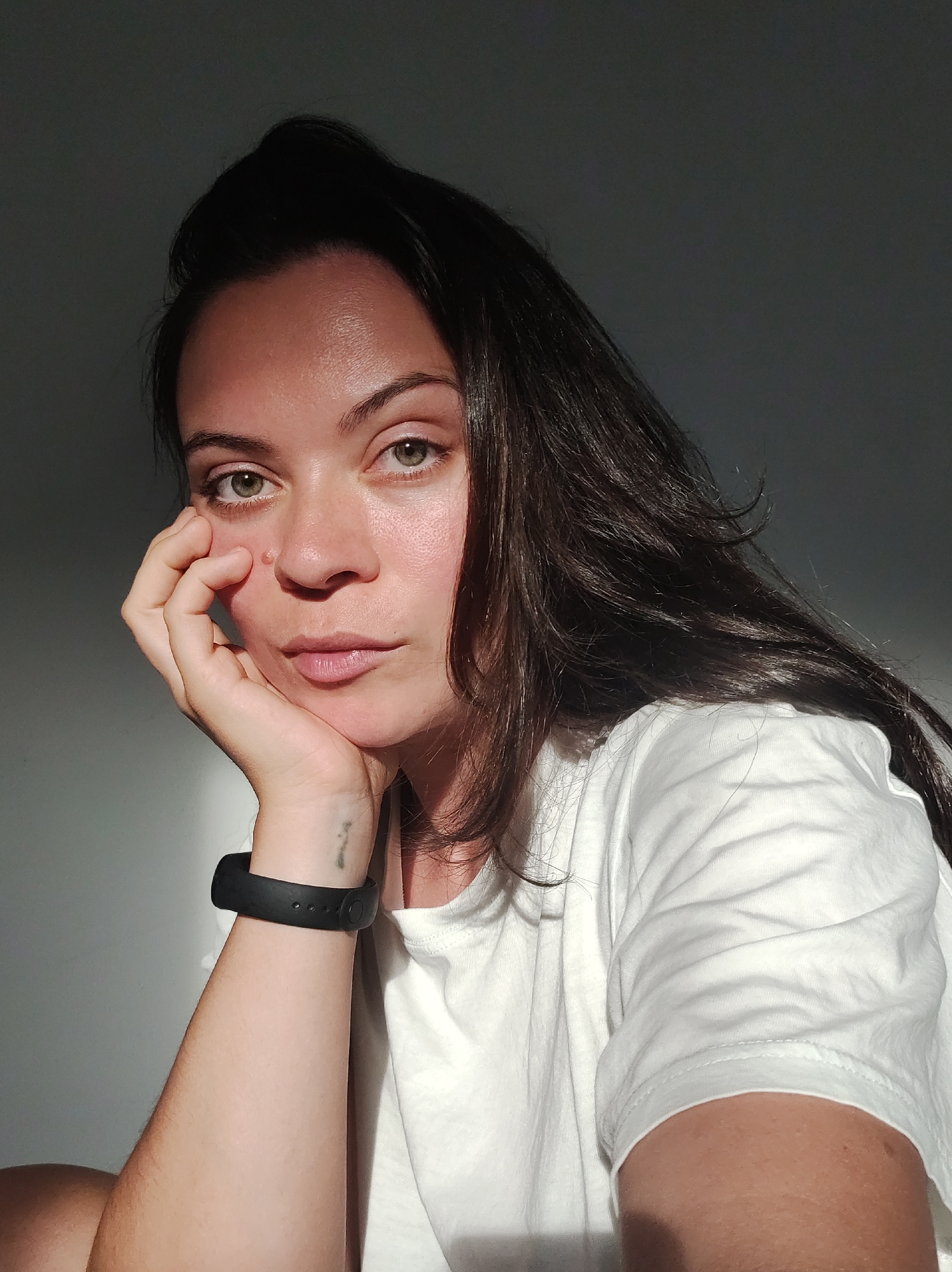 This is a close-up, medium-shot portrait of a young woman. She has long, dark hair and green eyes, and is looking directly at the camera with a neutral expression. Her right hand is propped under her chin, with her fingers resting on her cheek. She is wearing a white t-shirt and a black wristband on her left arm. A faint tattoo is visible on her left wrist.
The lighting suggests it is late afternoon, with warm sunlight casting shadows on her face and the wall behind her. The sun illuminates the left side of her face and highlights her hair. The setting appears to be indoors, possibly a room with a plain wall. The overall mood is contemplative and somewhat somber due to the lighting and her expression. There is no visible text in the image.