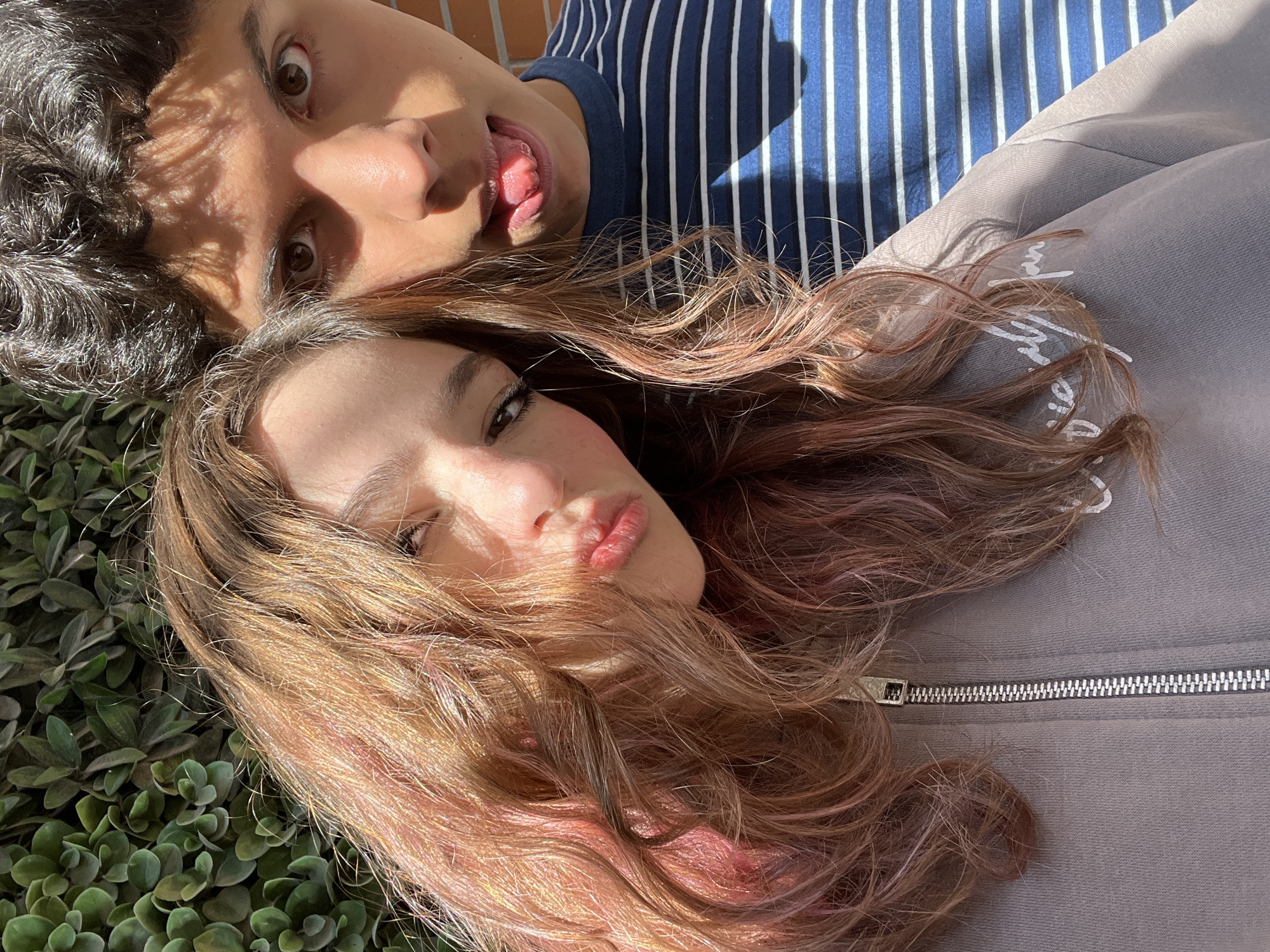 The image captures a close-up selfie of two young adults, a female and a male, located in Puebla, Mexico, under direct sunlight.
The female is positioned on the left, closer to the camera. She has long, wavy brown hair with prominent reddish-pink streaks, partially obscuring her face. Her head is tilted slightly, and she looks towards the camera with a pouty expression, her lips slightly pursed. She wears a light taupe or grey zippered hoodie with faint white text, possibly "Sincerely," visible on the left chest area.
The male is situated behind and to the right of the female, with only his upper face and torso visible. He has dark, curly hair and looks directly at the camera with wide eyes, his tongue partially extended in a playful manner. His face is marked by distinct shadows, consistent with foliage, across his forehead and eyes. He wears a dark blue and white horizontally striped shirt.
The background consists of a dense green foliage wall on the left and a red brick wall on the right. Strong overhead sunlight creates sharp shadows and highlights across both individuals and the environment.