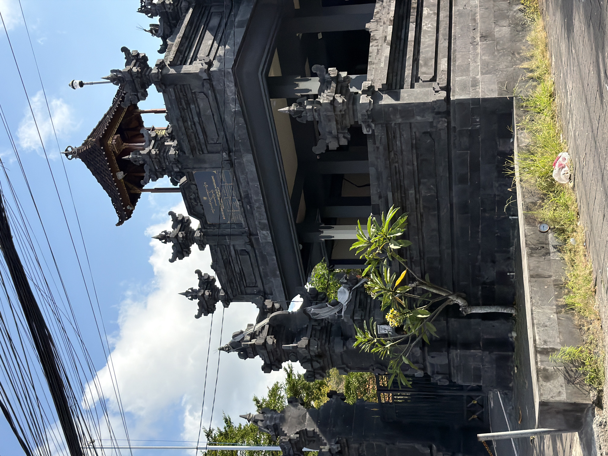 The image captures a detailed view of a traditional Balinese-style building, likely a temple gate or shrine, made of dark grey or black stone. The architecture is intricate, featuring multiple tiers, ornate carvings, and decorative finials, characteristic of structures found in Kuta Selatan, Indonesia. A prominent multi-tiered roof, crowned with a spire and a light fixture, dominates the upper part of the structure.
The sky is bright blue with scattered white clouds, indicating clear, sunny daylight. Numerous black electrical or communication wires crisscross the upper left portion of the frame against the sky. A small, leafy tree, possibly a frangipani with light-colored blooms, grows near the building's base on the right. The ground adjacent to the building is paved, with patches of green grass and weeds. A small, circular gauge or meter is visible on the ground near the grass, and a white plastic bag, possibly trash, lies nearby.
No specific activity or event is taking place; the scene is static. The architecture, intricate carvings, and the presence of a plaque with Balinese text serve as strong location cues for Bali.
A rectangular plaque on the building's facade prominently displays text. The top line reads: "ANUMERTA BHATARA GEDE MEGANTUNG." Below this, smaller, less distinct text is visible, with the word "PUJA" (worship) partially discernible, suggesting a dedication or purpose related to religious worship.