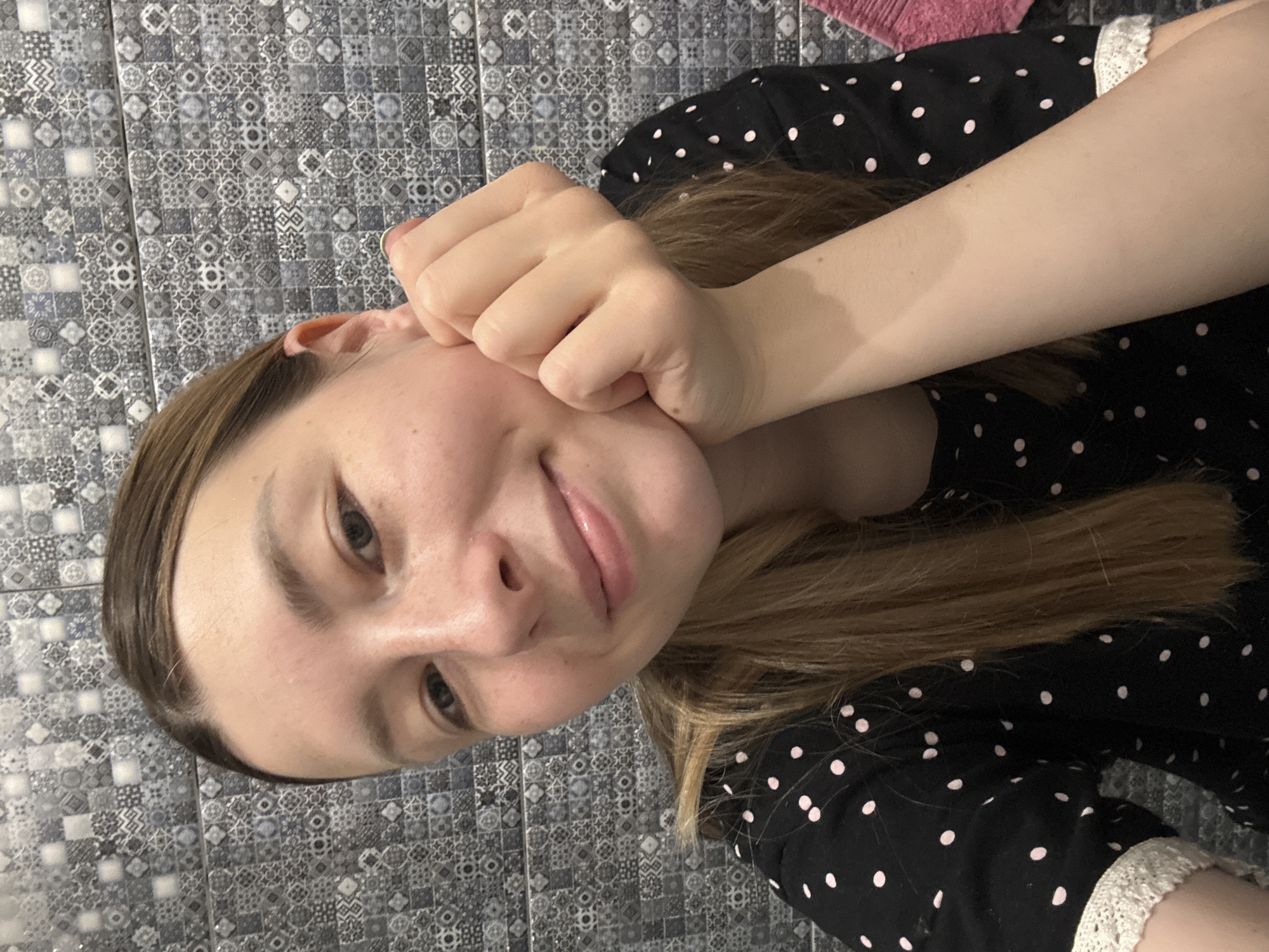 A woman is lying down in an interior space, likely a bathroom, given the patterned grey and white tile on the wall. She is wearing a black dress with white polka dots and lace trim on the sleeves. The woman's head rests on her left hand, which is clenched into a fist. Her blonde hair is partially visible. The scene shows no indication of being in Moscow, Russia.