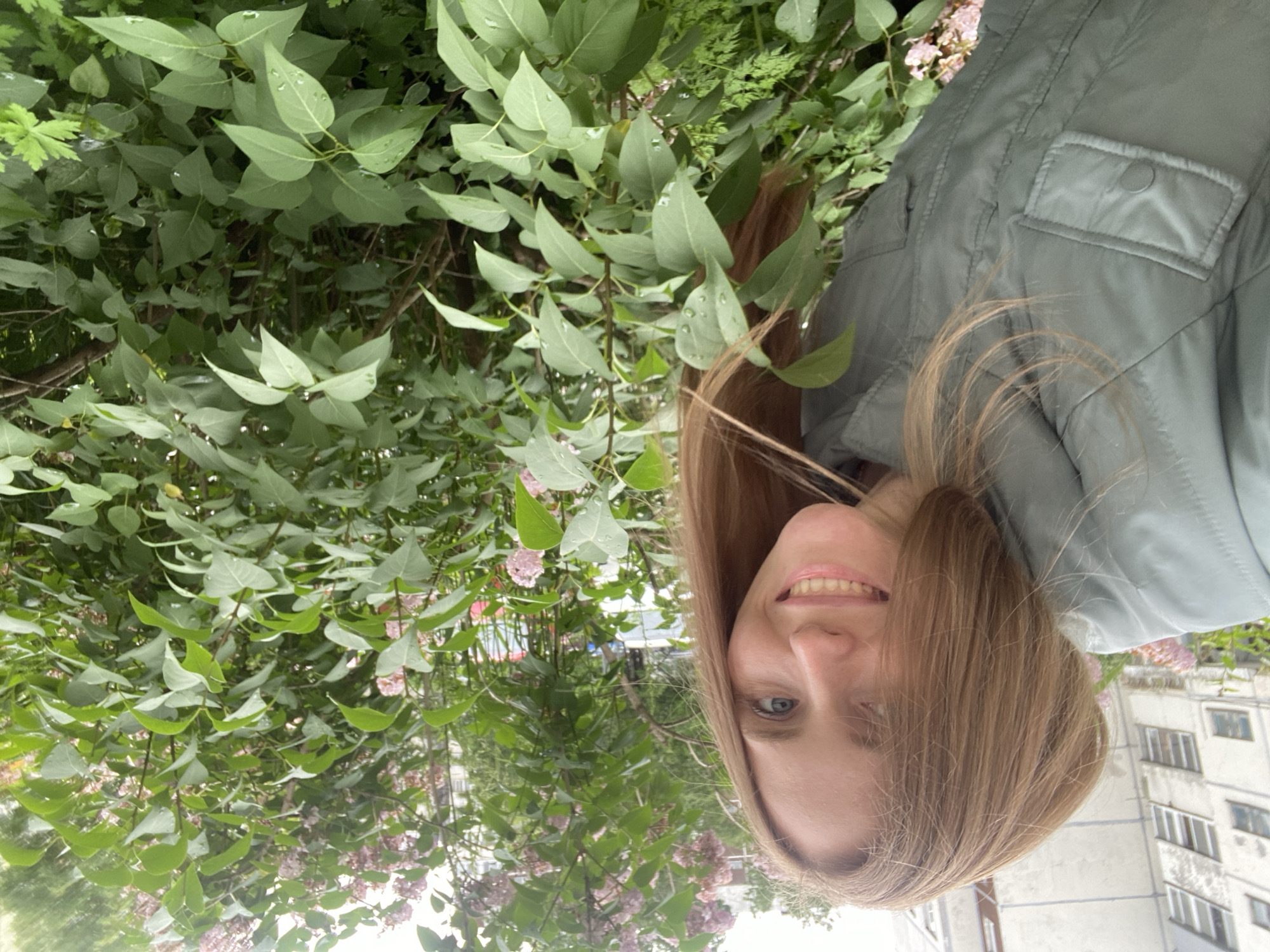 An outdoor, eye-level, medium shot shows a fair-skinned woman with long, light brown hair and blue eyes smiling at the camera. She is wearing a light green jacket. She is standing in front of a bush with green leaves and pink flowers. In the background, there is a building with white siding and windows. The scene appears to be in a residential area in Yuzhno-Sakhalinsk, Russia, as evidenced by the architectural style of the building and the presence of lilac bushes, which are common in Russian cities. The image has a shallow depth of field, with the woman in sharp focus and the background slightly blurred. The lighting is natural and diffused, suggesting an overcast day.
