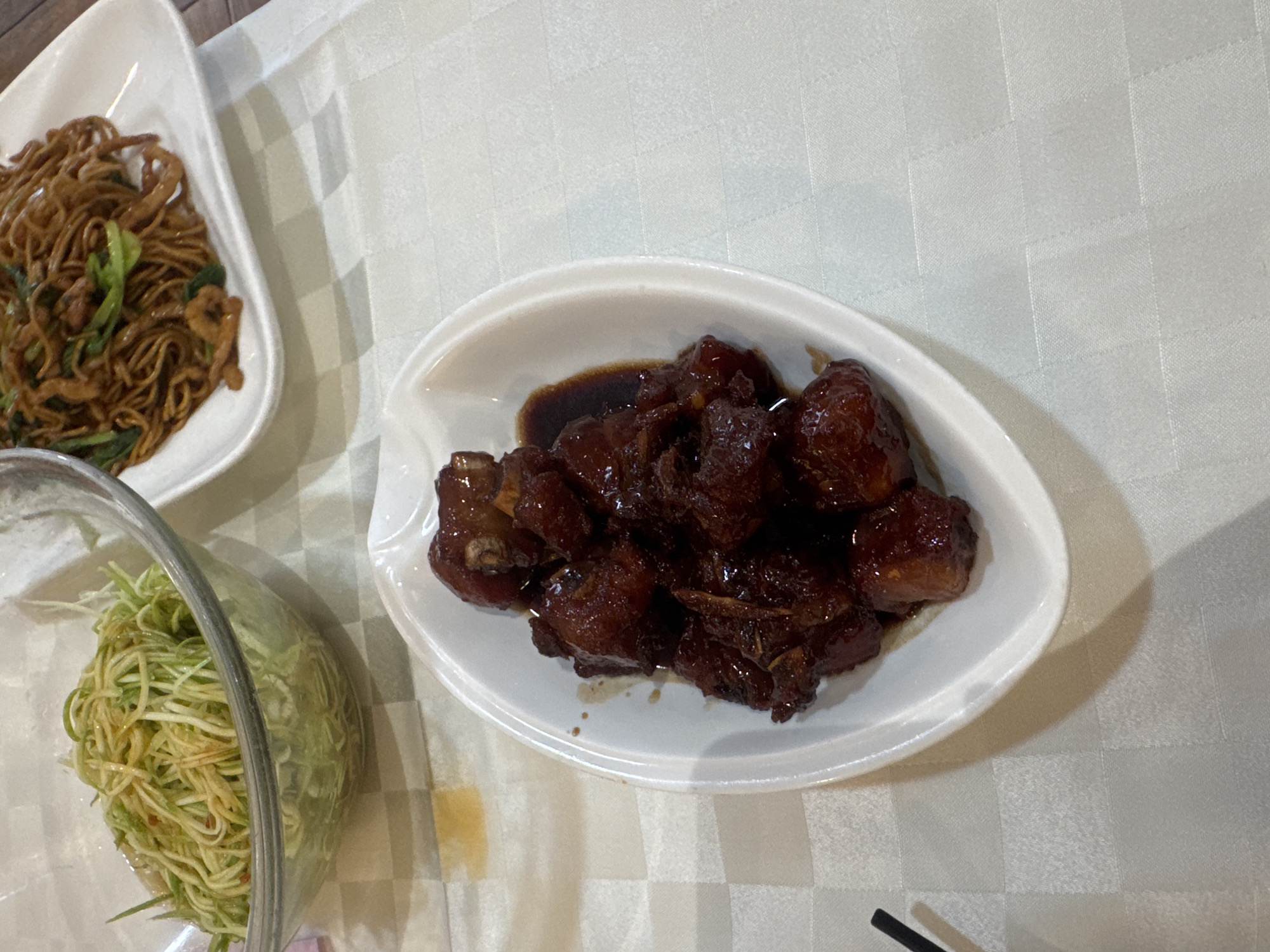 An overhead shot of a white tablecloth-covered table at a restaurant in Shanghai, China, shows three dishes of food. In the foreground, a white oval dish holds braised pork ribs in a dark, glossy sauce. To the left, a glass bowl contains a shredded vegetable salad with a light dressing. In the background, a white rectangular plate holds stir-fried noodles with vegetables and what appears to be meat. The setting appears to be a casual dining establishment.