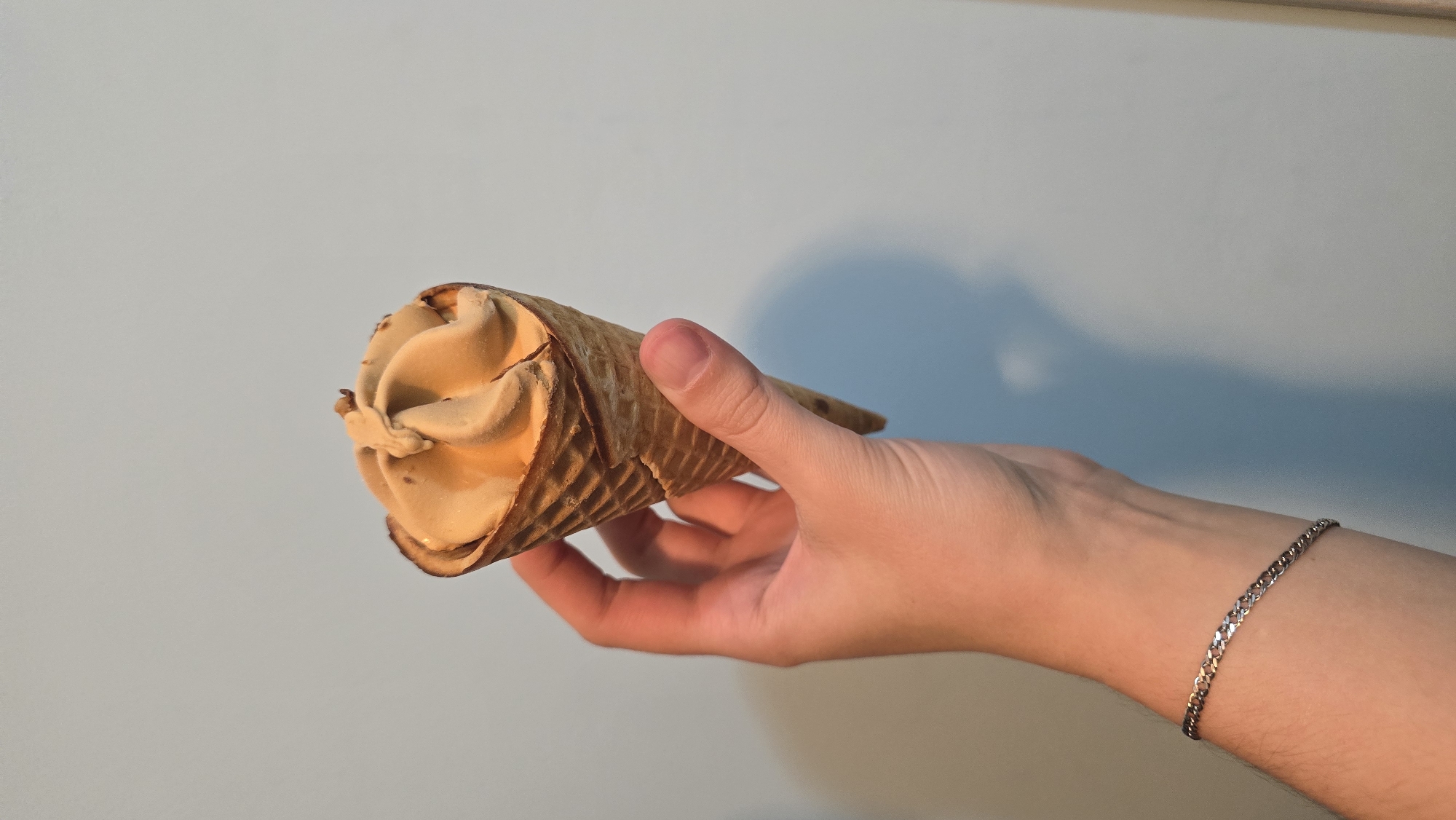 A person's hand holds a single, golden-brown ice cream cone with a swirl of light brown soft-serve ice cream. The ice cream appears to have some small dark flecks on top. The cone is a waffle cone with a diamond pattern. The hand is positioned in the center of the frame against a plain, light blue wall. The person is wearing a silver chain bracelet on their left wrist. The lighting creates a soft shadow on the wall behind the hand. There is no indication of the location being in a city.