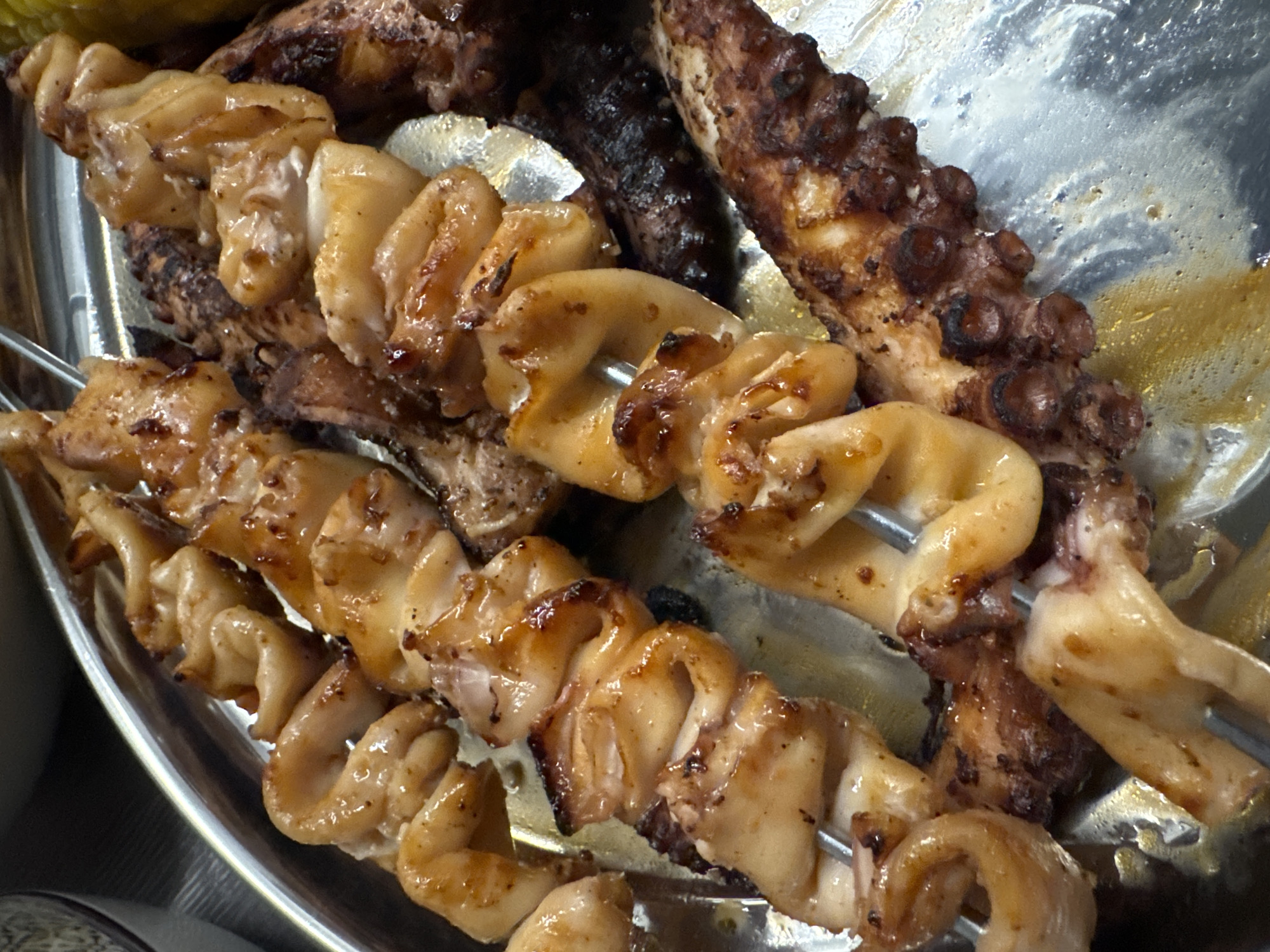 This image shows several skewers of grilled calamari and octopus tentacles arranged on a round, metallic platter. The calamari is sliced into rings and pieces, appearing glossy and slightly charred. The octopus tentacles are also grilled, with visible suckers and some darker, crispy edges. The platter is situated on a dark surface, possibly a table, and a sliver of a patterned bowl is visible in the upper left corner. The lighting is direct, highlighting the textures and glistening surfaces of the seafood. The setting appears to be an outdoor restaurant in Sustaš, Montenegro, characterized by the casual presentation of the food on a communal platter. There are no people or specific actions depicted, only the still arrangement of the grilled seafood.