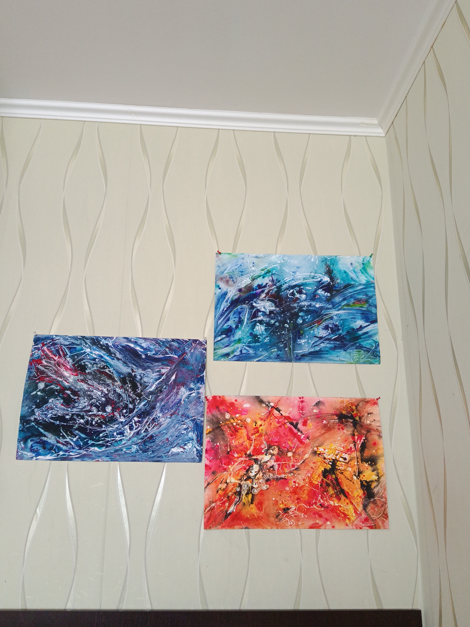The image shows three abstract paintings hanging on a wall. The wall has light beige wallpaper with a vertical wavy pattern and a white crown molding along the top. The paintings are rectangular and appear to be of different color schemes: one with blues and whites, one with lighter blues and some darker shades, and the third with oranges, reds, blacks, and yellows. The paintings are not framed, and the corners seem to be pinned or taped to the wall. A dark brown object is partially visible at the bottom of the image, likely a piece of furniture. The location is likely a residential space. There are no people in the image. The city and country are not apparent from this image.