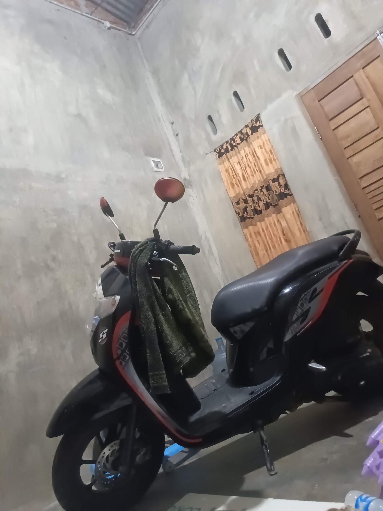 An indoor scene in what appears to be a garage or storage area, featuring a black and red scooter parked on a concrete floor. The scooter has a dark green cloth draped over its handlebars. The walls are unfinished concrete, and there is a wooden door with horizontal planks on the right side of the frame. A patterned orange and brown curtain hangs on the wall above and to the left of the door. The ceiling is exposed, showing some corrugated material and wiring. The lighting is dim, casting shadows on the floor and walls.