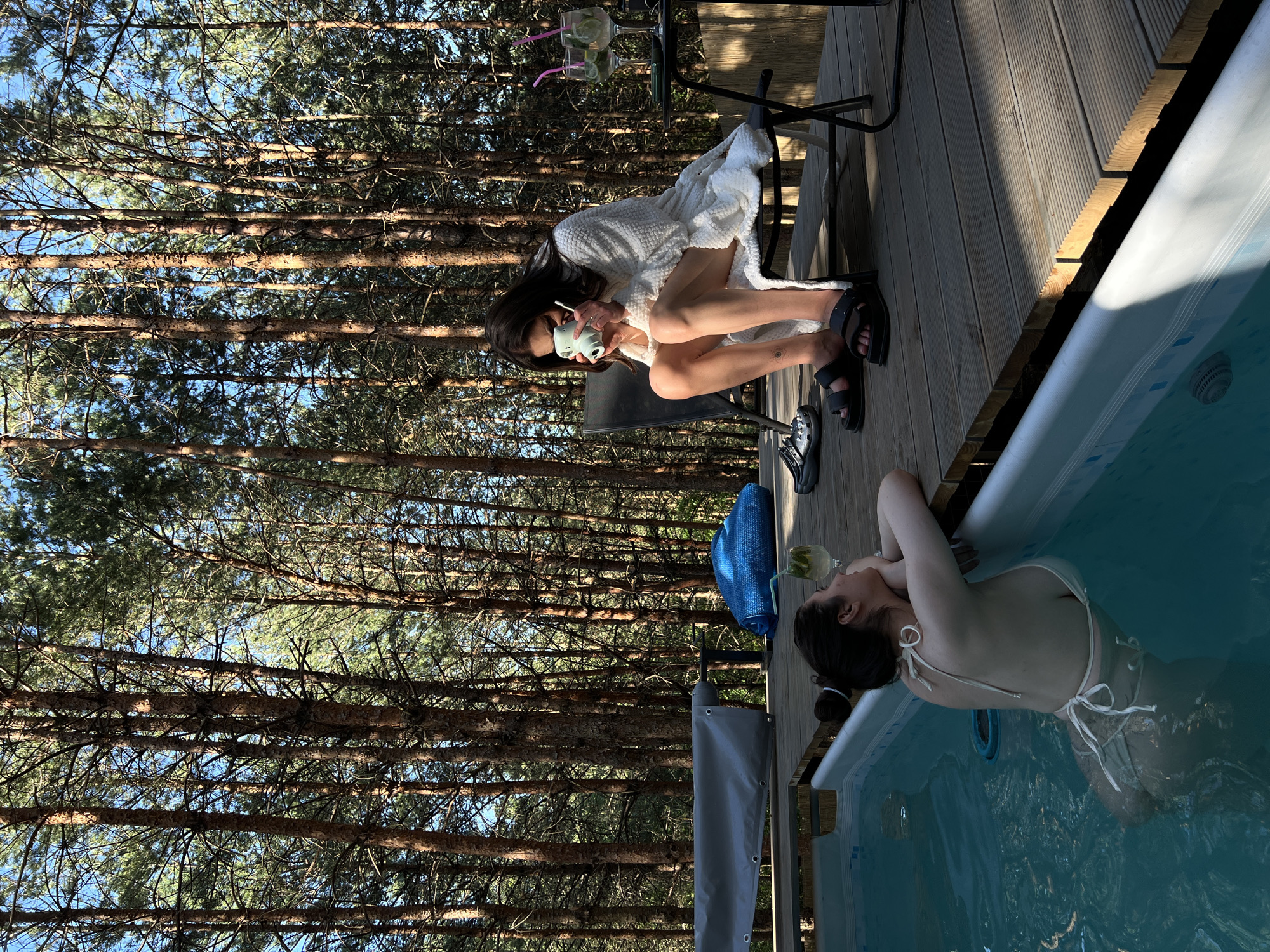 Two individuals are visible in an outdoor setting amidst a dense pine forest. In the foreground, a woman is partially submerged in a swimming pool, her back facing the camera. She is wearing a light-colored bikini and leaning on the edge of the pool. In the middle ground, a second woman is seated on a chair on a wooden deck, wearing a white bathrobe and holding a small camera to her face, as if taking a photo. Nearby, there is a small table with two drinks, and a blue towel lies on the deck. The environment suggests a private recreational area with a pool and seating, surrounded by tall pine trees. The location of Ivachiv Horishnii, Ukraine, is provided as context for this scene.