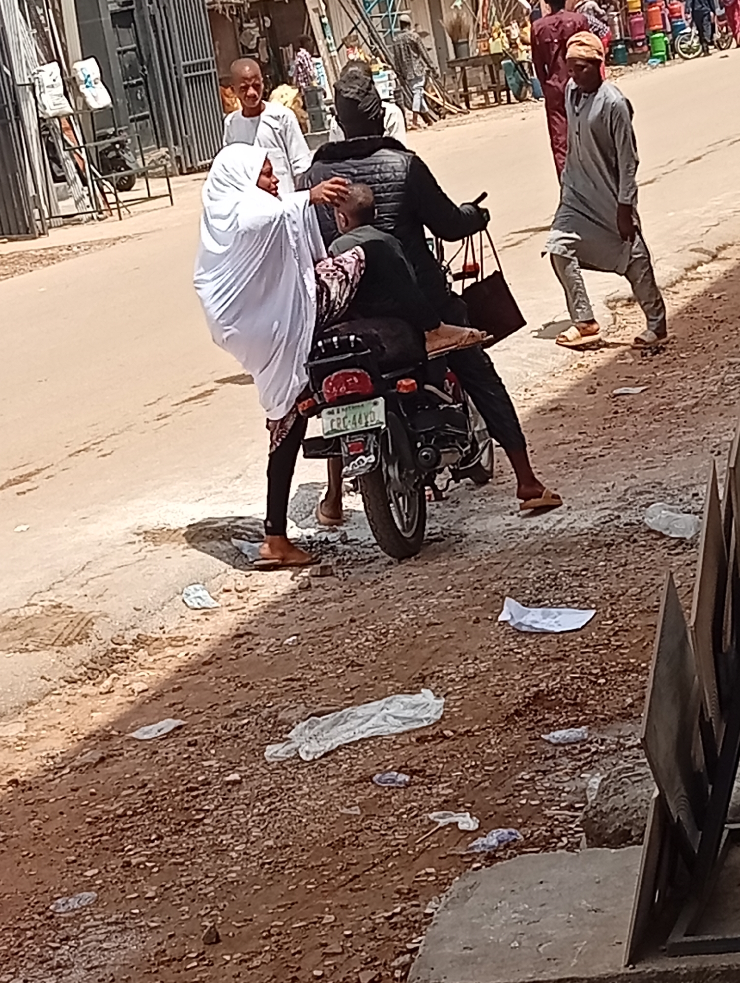 The image depicts a street scene in what is possibly Maiburiji, Nigeria. A motorcycle with three occupants is the focal point. The driver is wearing a black jacket and trousers, with a black head covering. The passenger directly behind the driver is wearing a black garment patterned with brown and white designs. The passenger behind that person is wearing a long white cloth covering their head and body. The motorcycle's license plate is partially visible, reading "CRC-44YD."
To the right of the motorcycle, a man in a gray robe and cap is walking on the sidewalk in a side profile. In the background, other figures are also present, and what looks like a market area with colorful goods on display. The ground is a mix of paved road and unpaved, gravelly area with scattered trash, including plastic bags. A gate or fencing structure is visible on the left side of the frame.
