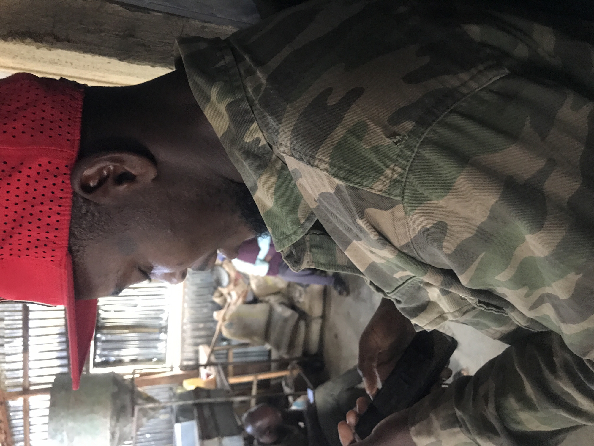 The image depicts a close-up view of a young Black man wearing a red baseball cap and a camouflage jacket, holding a smartphone in his hands. The man's head is tilted downwards as he appears to be focused on the device. In the background, the scene suggests an indoor environment that might be a storage area or workshop, with corrugated metal walls and ceiling. Several sacks are stacked in the background, and other individuals are partially visible in the blurred distance. The lighting is somewhat dim, with light filtering in from an unseen source, casting shadows and highlighting certain areas. The man's skin tone is dark. The overall impression is one of a casual moment captured in what seems to be a functional, perhaps industrial or rural setting.