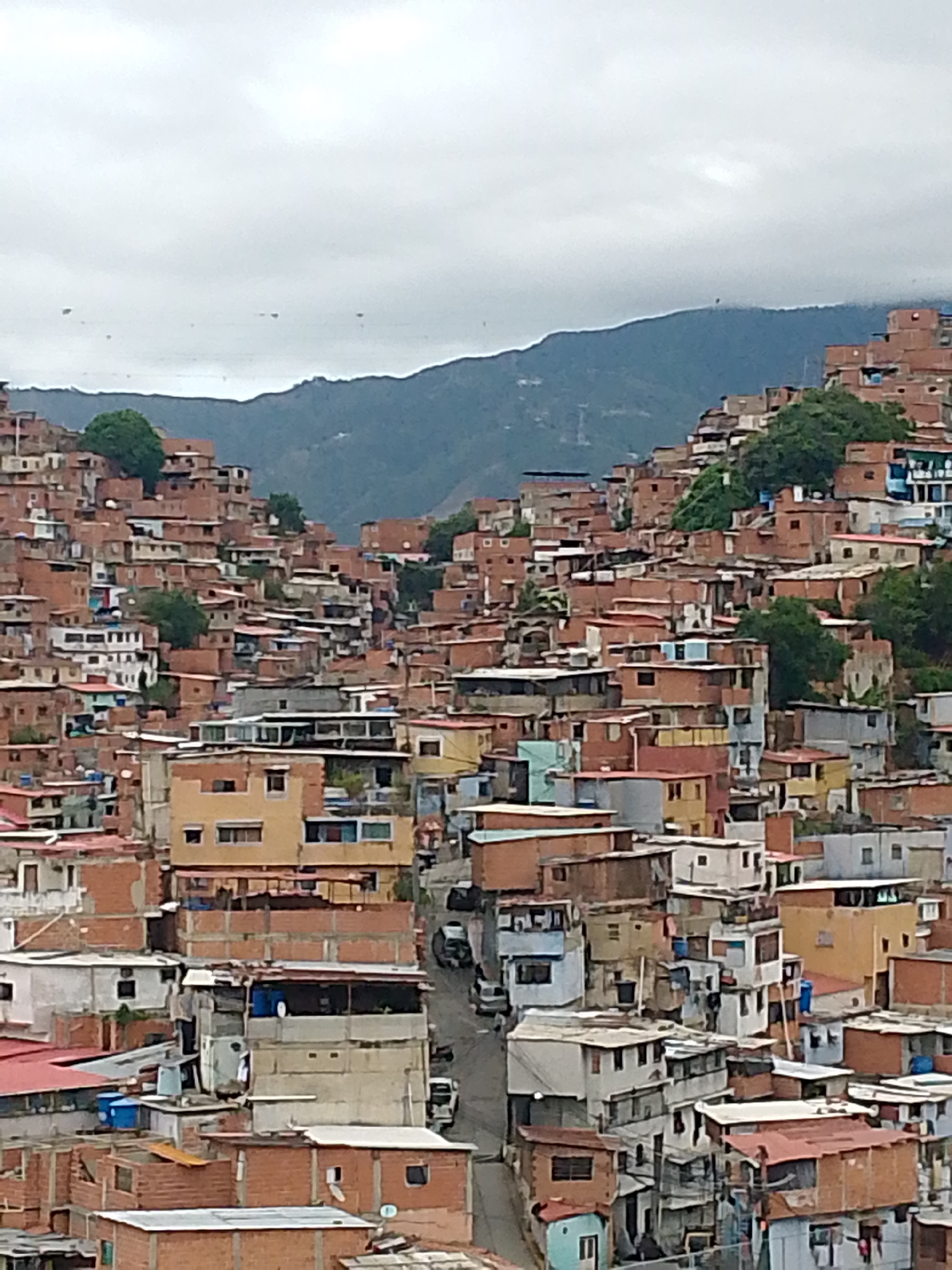 The image shows a densely populated urban area in Caracas, Venezuela, likely a shantytown or favela. The scene is dominated by tightly packed, multi-story buildings constructed primarily of brick and concrete, with various paint colors and materials. A road winds through the center of the built environment. In the background, a mountain range is visible under an overcast sky. The overall environment appears impoverished and informal.