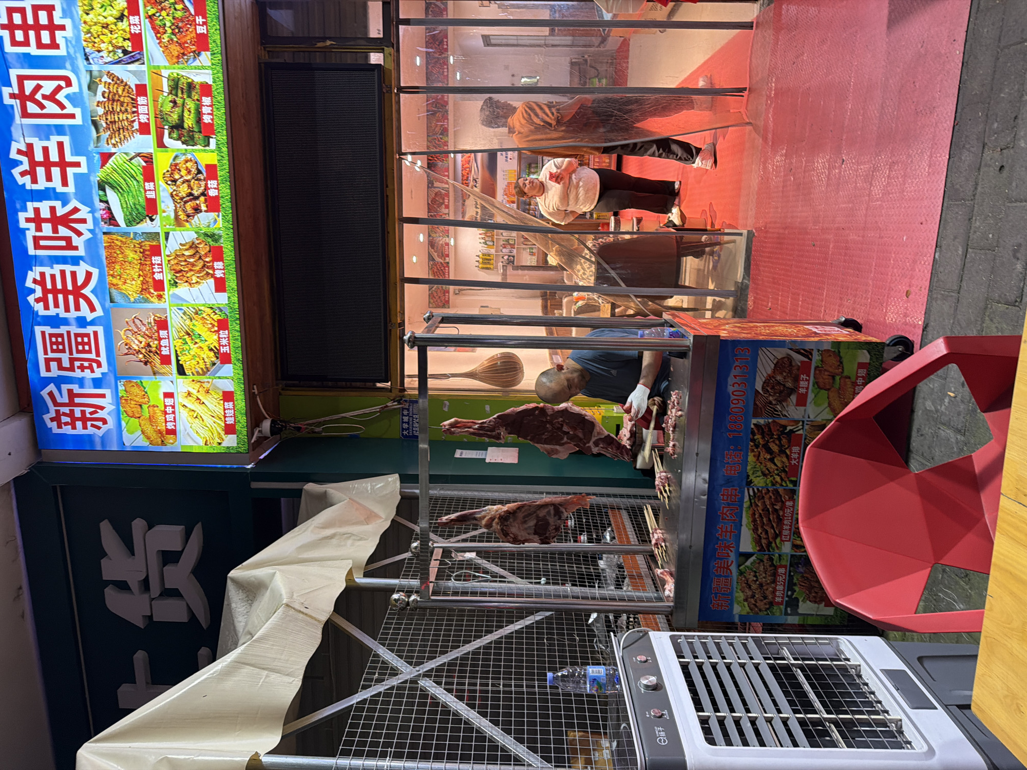 Here's a description of the scene:
The image shows a street food stall in the Chongqing High-tech Zone, China. The stall is illuminated by a bright sign with Chinese characters advertising Xinjiang-style lamb skewers. The sign also lists other grilled items such as chicken wings, squid, mushrooms, and vegetables.
Inside the stall, a man wearing gloves is preparing lamb skewers. Two large pieces of raw lamb are hanging from metal racks. A metal wireframe structure surrounds the food preparation area. A small electric grill is positioned nearby, ready for cooking.
Customers are visible through a glass paneling of the food stall. A red plastic chair is positioned on the right side of the image, facing the direction of the stall.