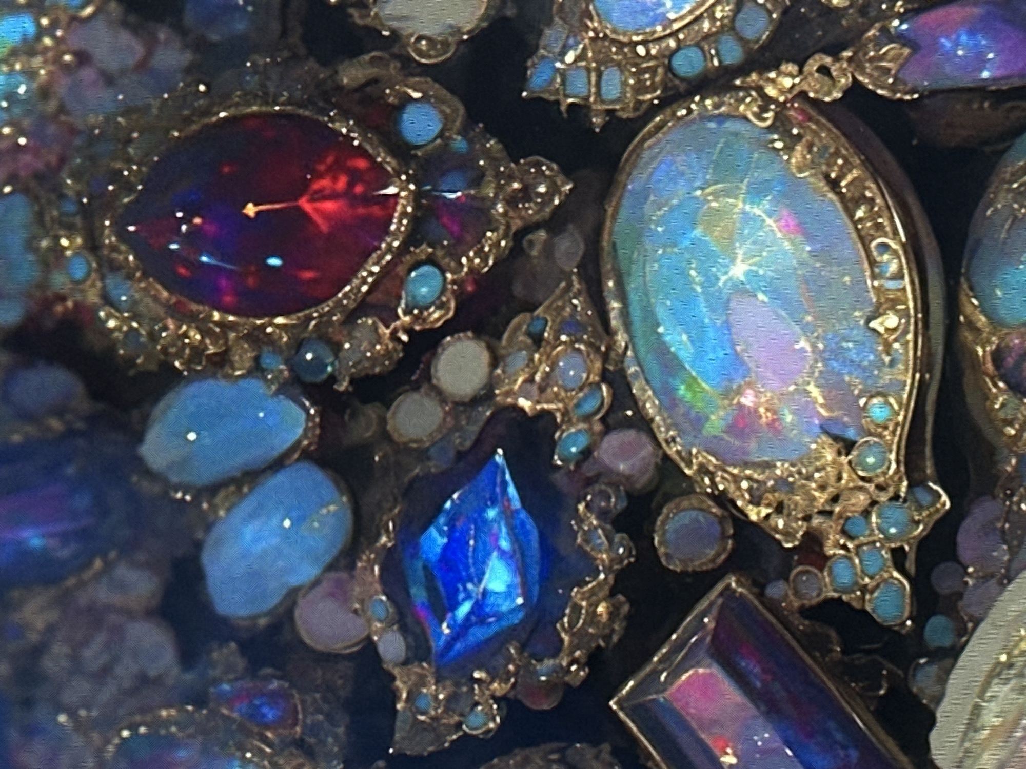 The image is a close-up, overhead shot of an assortment of ornate jewelry pieces, possibly part of a collection or display. The pieces are characterized by large, multifaceted gemstones in vibrant shades of blue, red, and iridescent opal. These stones are set in intricate gold metalwork, often accented with smaller turquoise or blue cabochons. The lighting appears to be artificial, creating highlights and reflections on the polished surfaces of the jewelry. The background is dark and indistinct, focusing all attention on the detailed craftsmanship and rich colors of the jewels. The overall impression is one of opulence and luxury.