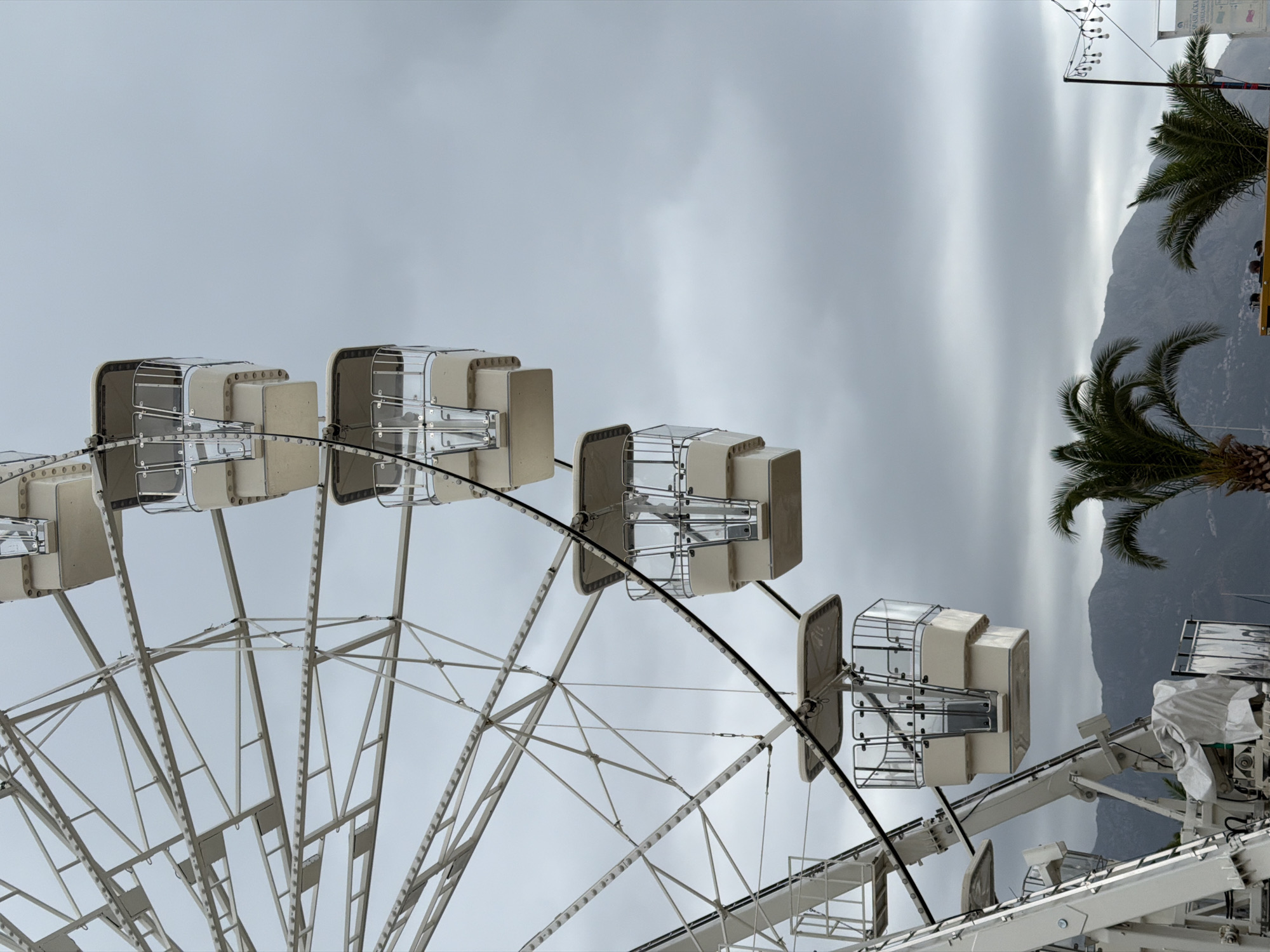 The image captures a close-up view of a white Ferris wheel, with several gondolas visible against a cloudy sky. The Ferris wheel structure is intricate, composed of numerous white metal beams and supports. Some gondolas are suspended from a thick black cable that runs parallel to the wheel's spokes. The gondolas themselves are boxy and have transparent sides, allowing a glimpse of their interior. In the background, a range of muted, mountainous terrain is visible, suggesting an outdoor setting. Palm trees are also present, with their fronds reaching towards the sky.
The scene is located in Budva, Montenegro, likely in a tourist area due to the presence of the Ferris wheel and palm trees. The overall atmosphere is subdued, owing to the overcast weather. No people are explicitly visible interacting with the Ferris wheel or its components. The image focuses on the mechanical and structural aspects of the Ferris wheel, with the natural landscape serving as a backdrop.