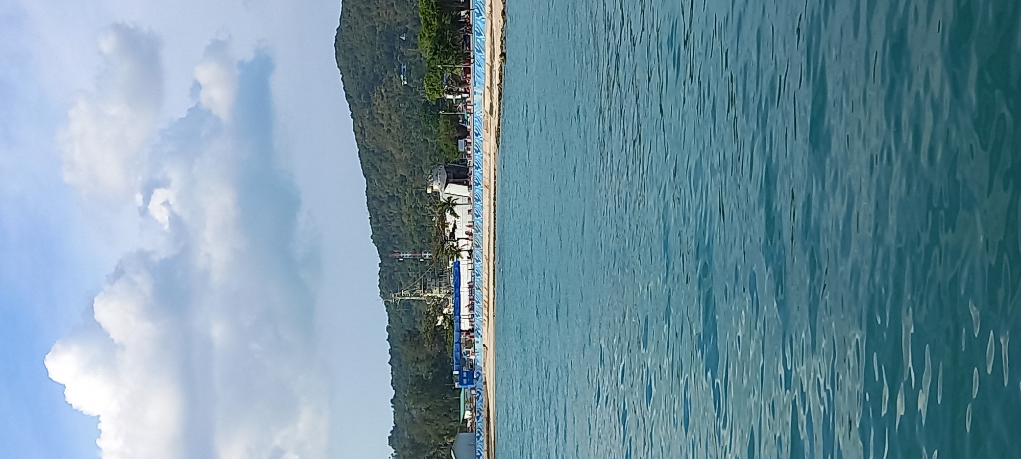 The image displays a vibrant coastal landscape, oriented vertically. On the right side, a vast expanse of calm, turquoise to blue-green ocean water dominates, featuring numerous ripples and gentle undulations on its surface.
The left half of the image transitions from the sky at the far left to a mountainous coastline. The far left edge shows a bright blue sky dotted with scattered white and light grey cumulus clouds. Below the sky, a steep, verdant mountain, densely covered in tropical green foliage, forms a significant part of the landscape.
At the base of this mountain, along the water's edge, a developed coastline is visible. It features several structures, including white and darker buildings, utility poles with wires, and prominent palm trees, suggesting a populated area. A distinctive long wall or barrier, adorned with a blue and white decorative pattern, runs parallel to the water, likely part of a pier or waterfront promenade.
No specific human activity or identifiable individuals are discernible in the frame. The scene captures a peaceful daytime setting, indicated by the bright, natural illumination and clear visibility. The partly cloudy weather conditions are evident from the sky. The lush tropical mountain, palm trees, and the clear blue water strongly suggest a tropical location, aligning with the context of Thong Sala, Thailand. There is no visible text in the image.