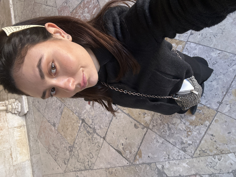 A female subject is depicted in a high-angle selfie, looking directly at the camera. She has dark brown hair, styled with a light-colored, studded hair clip on her right side. She is wearing a dark, long-sleeved garment, possibly a coat, and carries a small, metallic, chainmail-style handbag with a long silver-toned chain strap, worn across her body. She stands on a patterned floor composed of irregular, light-colored stone tiles in shades of grey and beige. In the background, a light-colored stone wall displays an ornate, intricately carved architectural element, identifiable as the base of a column or pilaster in the Manueline Gothic style. The specific architectural details and flooring indicate the location is within the Jerónimos Monastery (Mosteiro dos Jerónimos) in Belém, Lisbon, Portugal, likely within one of its cloisters or arcades.