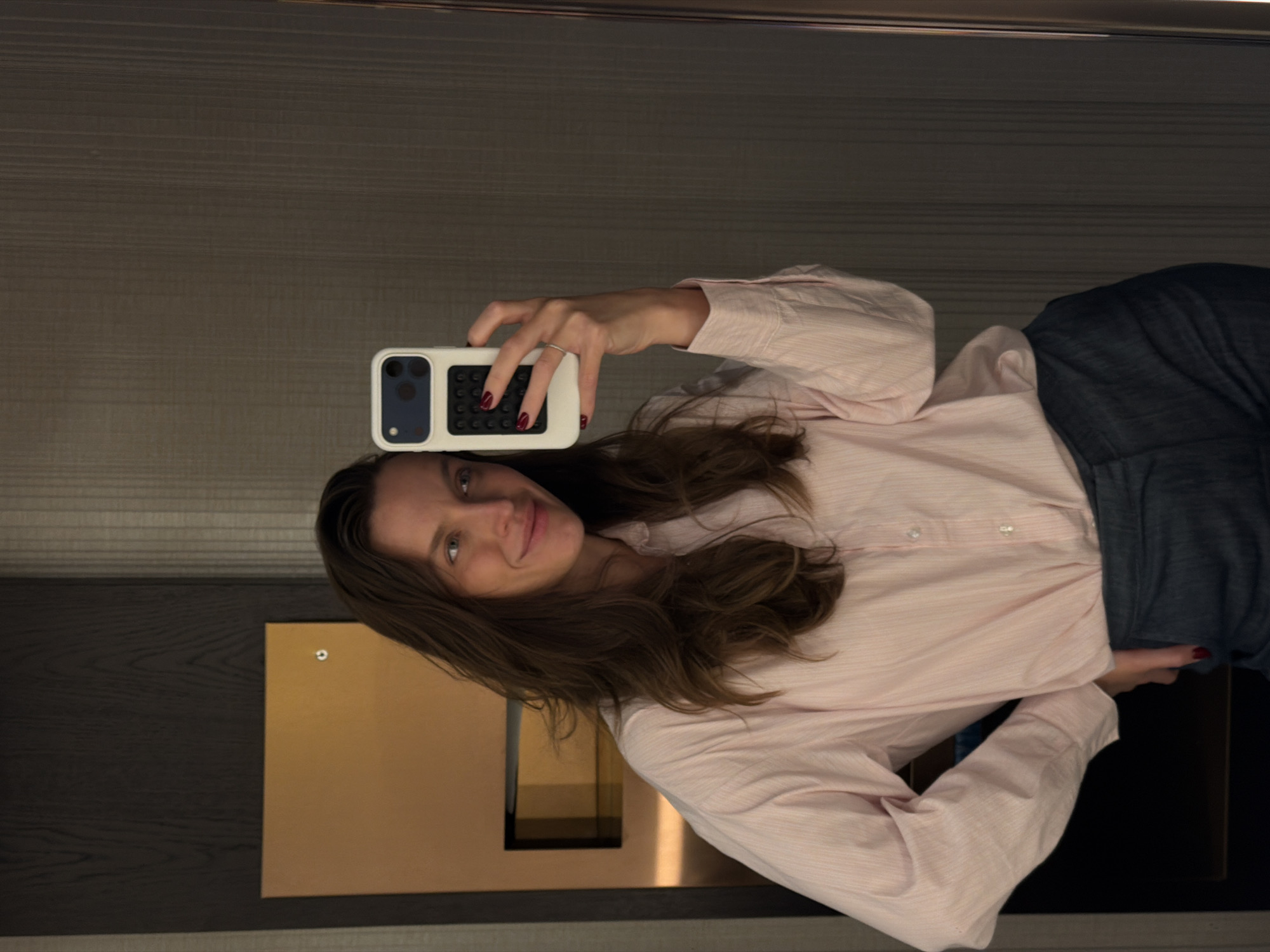 A woman is taking a selfie in front of a mirror. She is wearing a light pink striped shirt and dark jeans. She has brown hair and is smiling slightly. She is holding a smartphone with a white case in her right hand. The background shows a modern interior with a dark wooden wall and a gold-colored panel. The location is possibly in Budva, Montenegro, but this cannot be confirmed from the image alone.