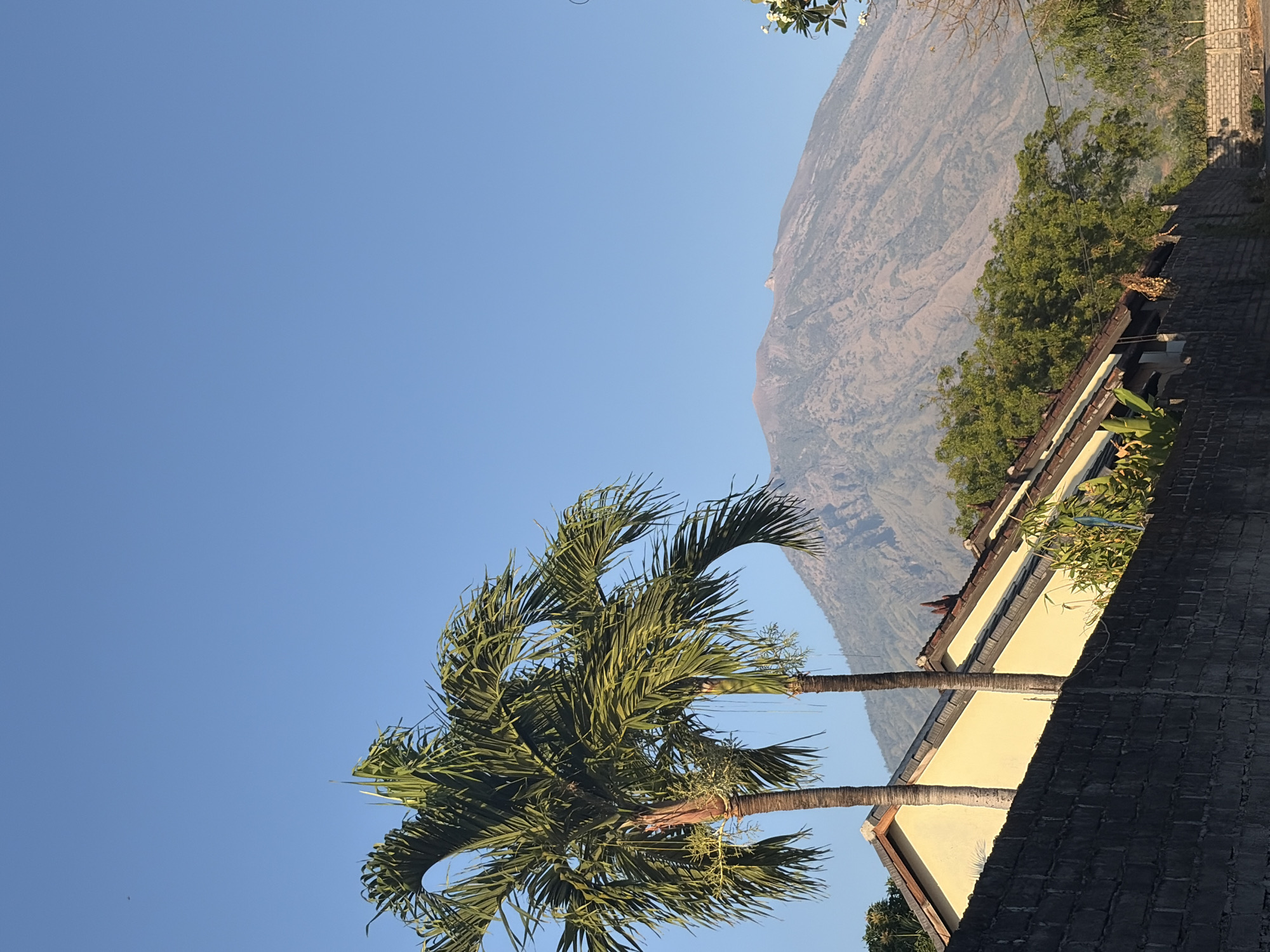 This is a landscape photograph taken in Kubu, Indonesia, under a clear blue sky. In the foreground, a dark brick wall runs horizontally across the lower portion of the frame. Behind the wall, a light yellow house with a brown tiled roof is visible. Two palm trees stand in front of the house, with their fronds extending towards the sky. In the background, a large, dormant volcano dominates the horizon, its slopes showing signs of dried vegetation and subtle contour lines. The overall impression is one of a serene, rural setting.