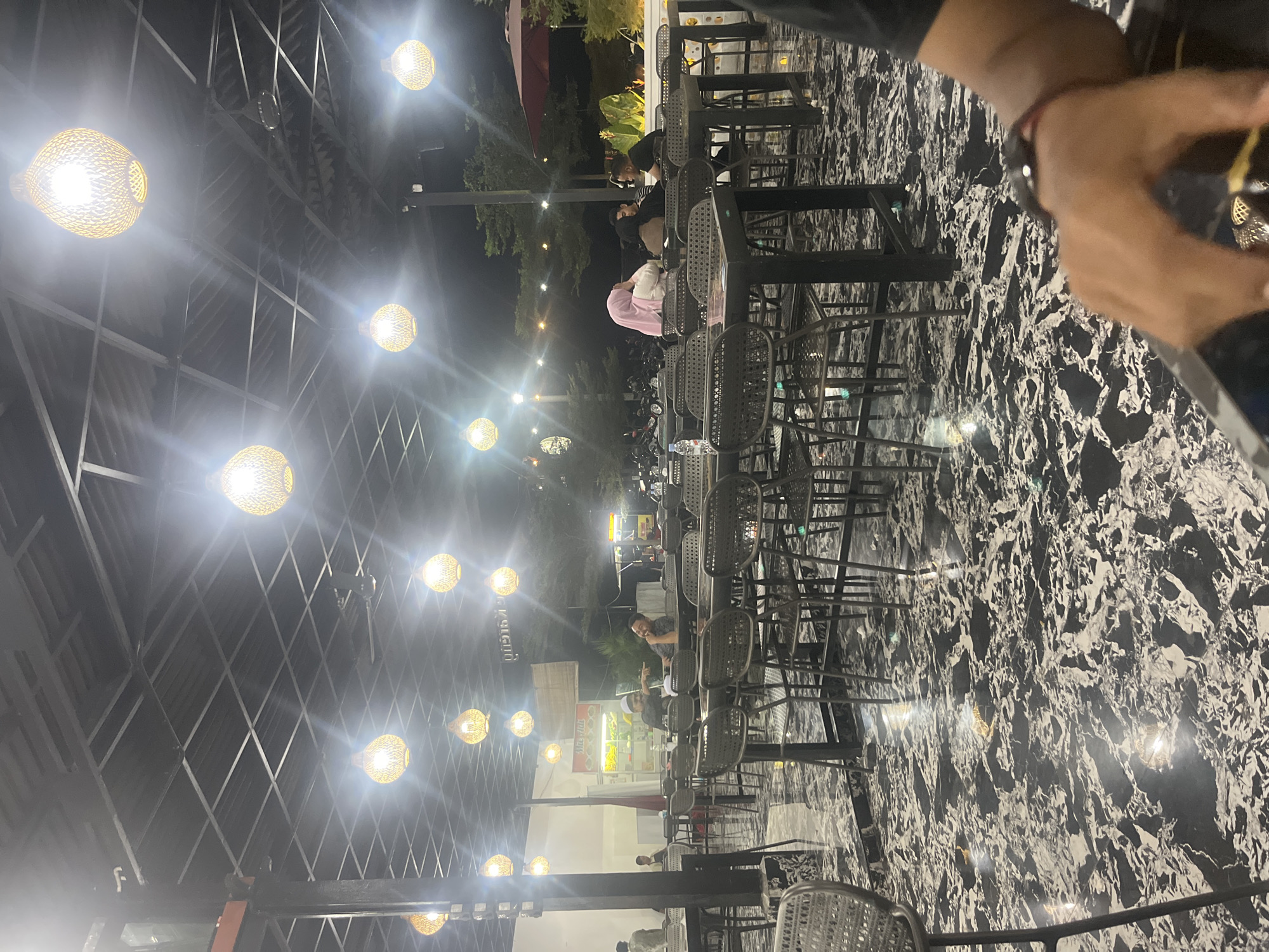 This image captures a nighttime scene at an open-air restaurant in Glameunasah Baro, Indonesia. The overhead structure consists of dark, exposed metal beams and a roof, from which numerous spherical woven light fixtures are suspended, providing warm illumination. A ceiling fan is also visible within the structure. The floor is a highly reflective black surface with irregular white patterns, resembling polished stone, reflecting multiple light sources.
Numerous patrons are seated at dark rectangular tables with matching dark, high-backed chairs. In the mid-ground, four individuals are seated at one table: one wearing a pink hijab and white top is seated with their back partially to the camera, while another in a striped shirt, a third in a dark top (partially obscured), and a male in a dark t-shirt are visible. A plastic water bottle rests on their table. Further to the left in the background, two men are interacting; one, wearing a white head covering and dark shirt, points towards the second man, also in a dark shirt, who is laughing. Additional indistinct patrons are scattered throughout the seating area.
In the background, a food vendor stall with a white facade and a sign displaying partial text "Dusun" and "Sate Hut" is visible to the left. To the right, a large red outdoor parasol is present. Beyond the restaurant area, parked motorcycles and trees are discernible against the dark night sky.
In the immediate foreground on the lower right, a person's right arm and hand are visible, holding a dark object, likely a mobile phone. A black wrist watch with a red band is worn. This perspective indicates the image was captured by the individual in the foreground.