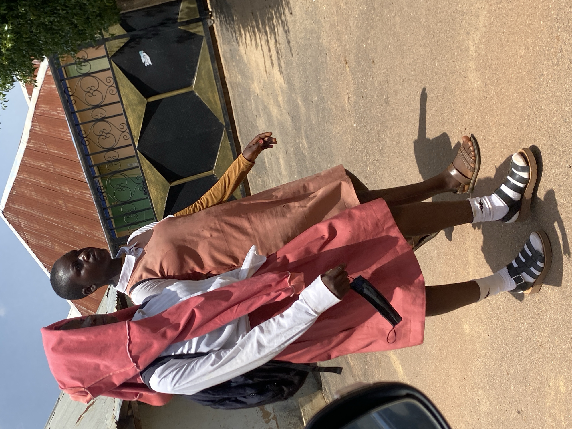 Two individuals, appearing to be young women or girls, walk on a paved surface in Bar Jirgi Summa, Nigeria. The individual on the left wears a white long-sleeved shirt, a pink skirt, and a pink head covering. They are also carrying a black backpack and holding a black face mask. The individual on the right wears an orange dress with a brown long-sleeved undergarment. Both are wearing sandals. Behind them is a building with a red corrugated metal roof and a black and gold decorative gate with ornate scrollwork. The ground is a light-colored asphalt or concrete with shadows cast from an unseen overhead object. The sky is visible and appears to be clear and blue.