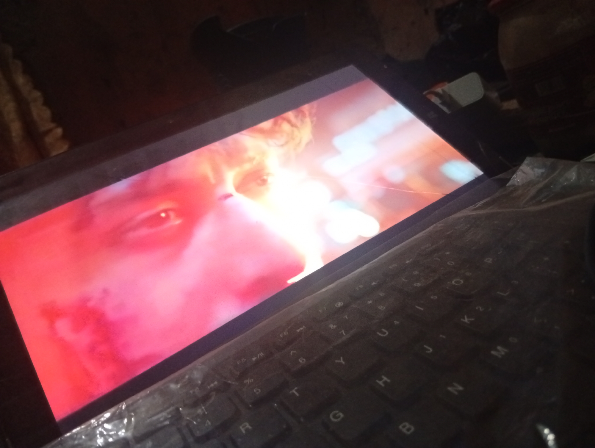 A close-up, angled shot shows a laptop screen displaying a close-up of a person's face, predominantly in shades of red and pink. The person's right eye is visible, looking directly at the camera, with a hint of blonde hair. The screen is positioned above a black keyboard, which is partially covered by a clear plastic sheet. The keyboard shows visible letters and symbols, and the overall scene is dimly lit. The background is dark and out of focus, with some blurred shapes suggesting a cluttered environment. The image is taken from a slightly elevated perspective.